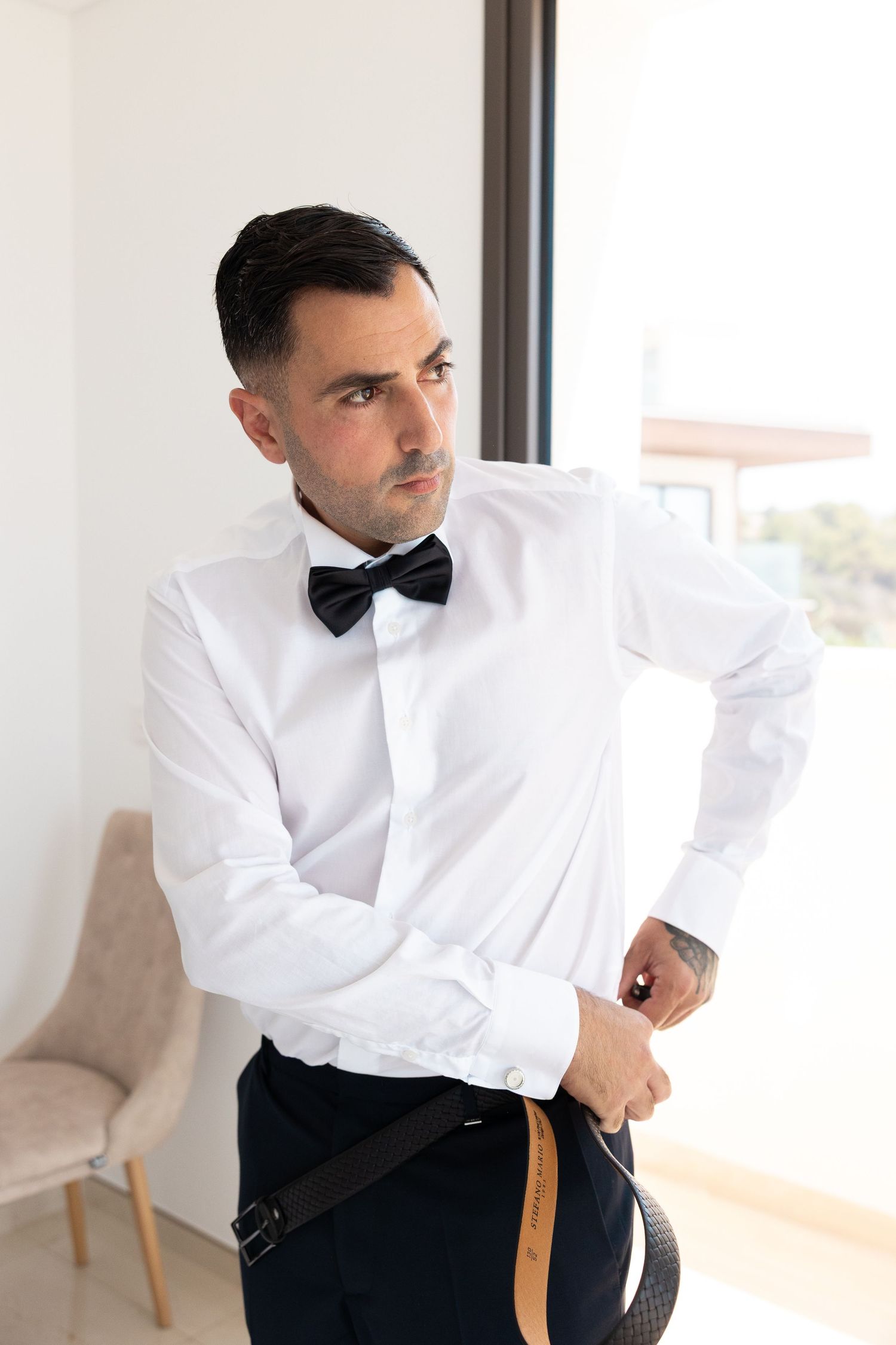 Person in formal attire with white dress shirt and black bow tie leans against furniture in bright interior space.