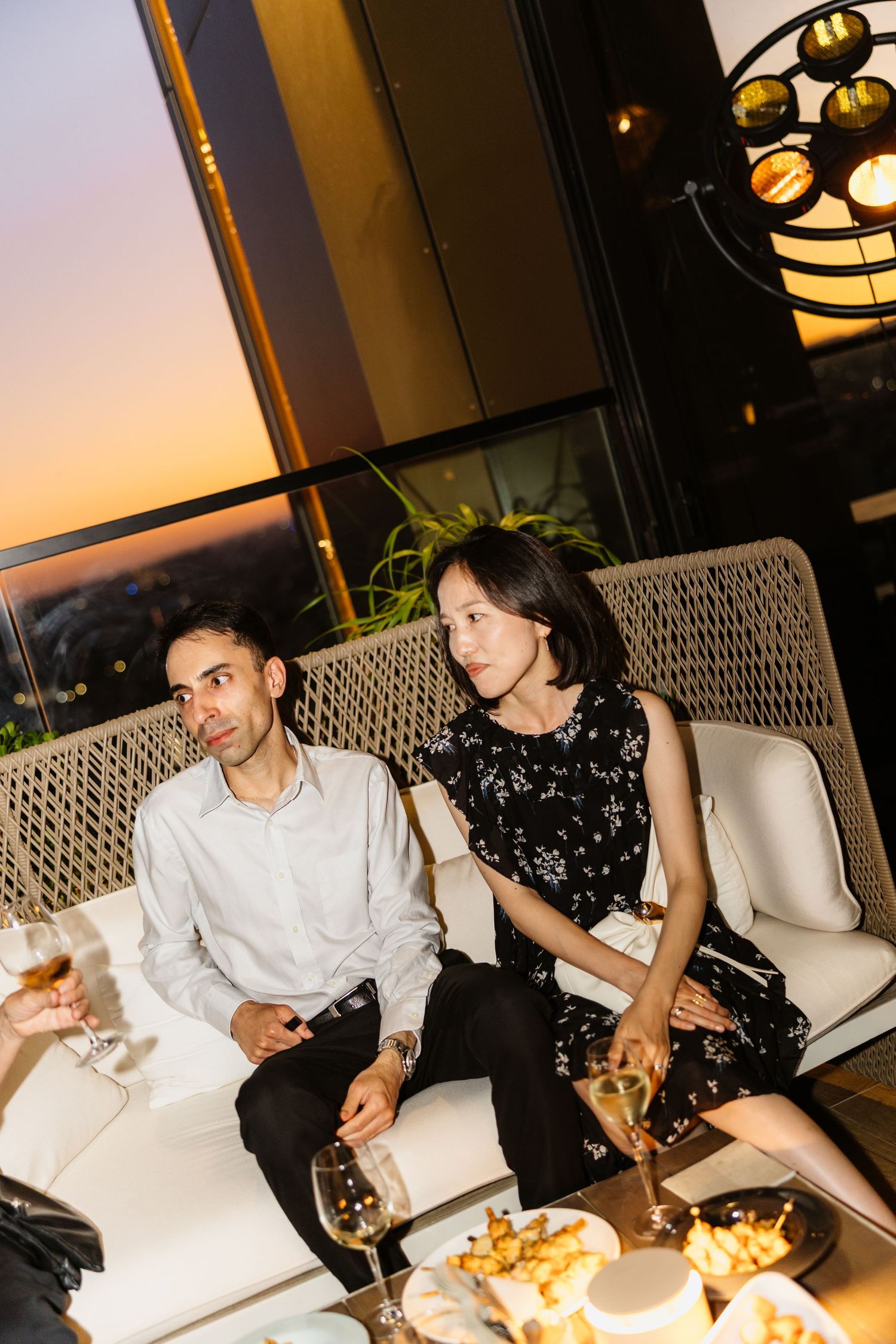 Elegant couple lounging on white outdoor sofa at sunset with cocktails and appetizers.
