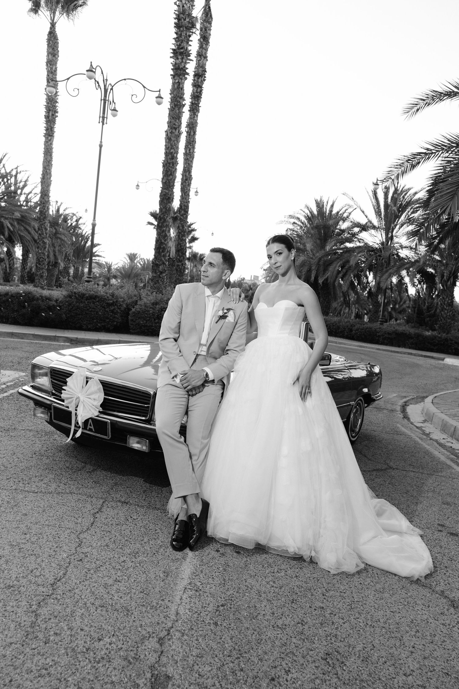 A bride and groom pose with a classic car on a palm tree-lined street in black and white photography.