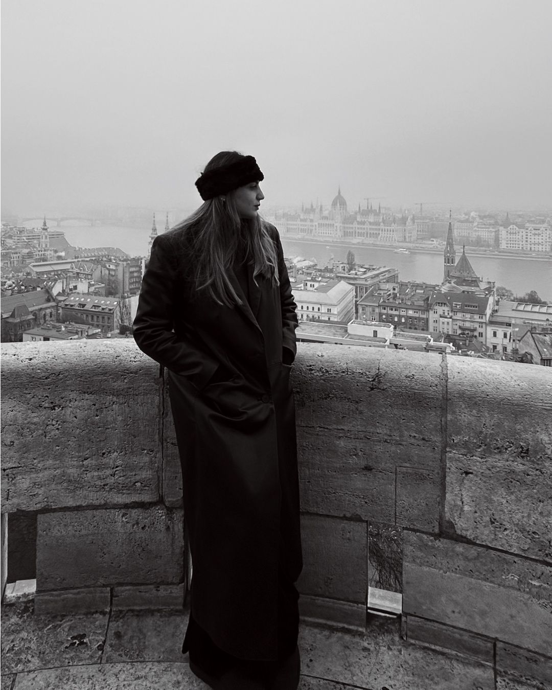 Black and white photo of person in long coat overlooking Budapest cityscape.