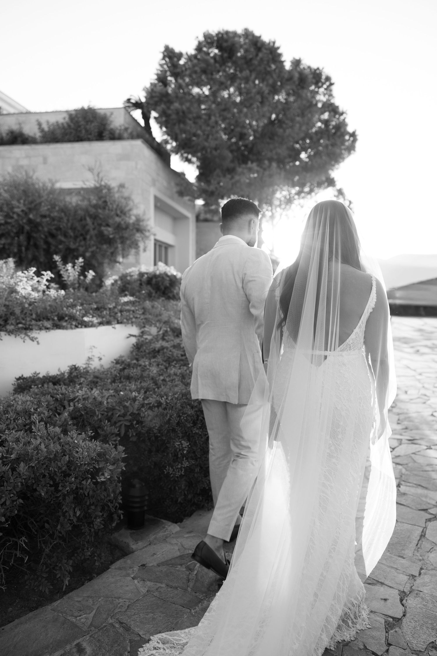 Black and white photo of bride in lace gown and groom walking away on stone path.