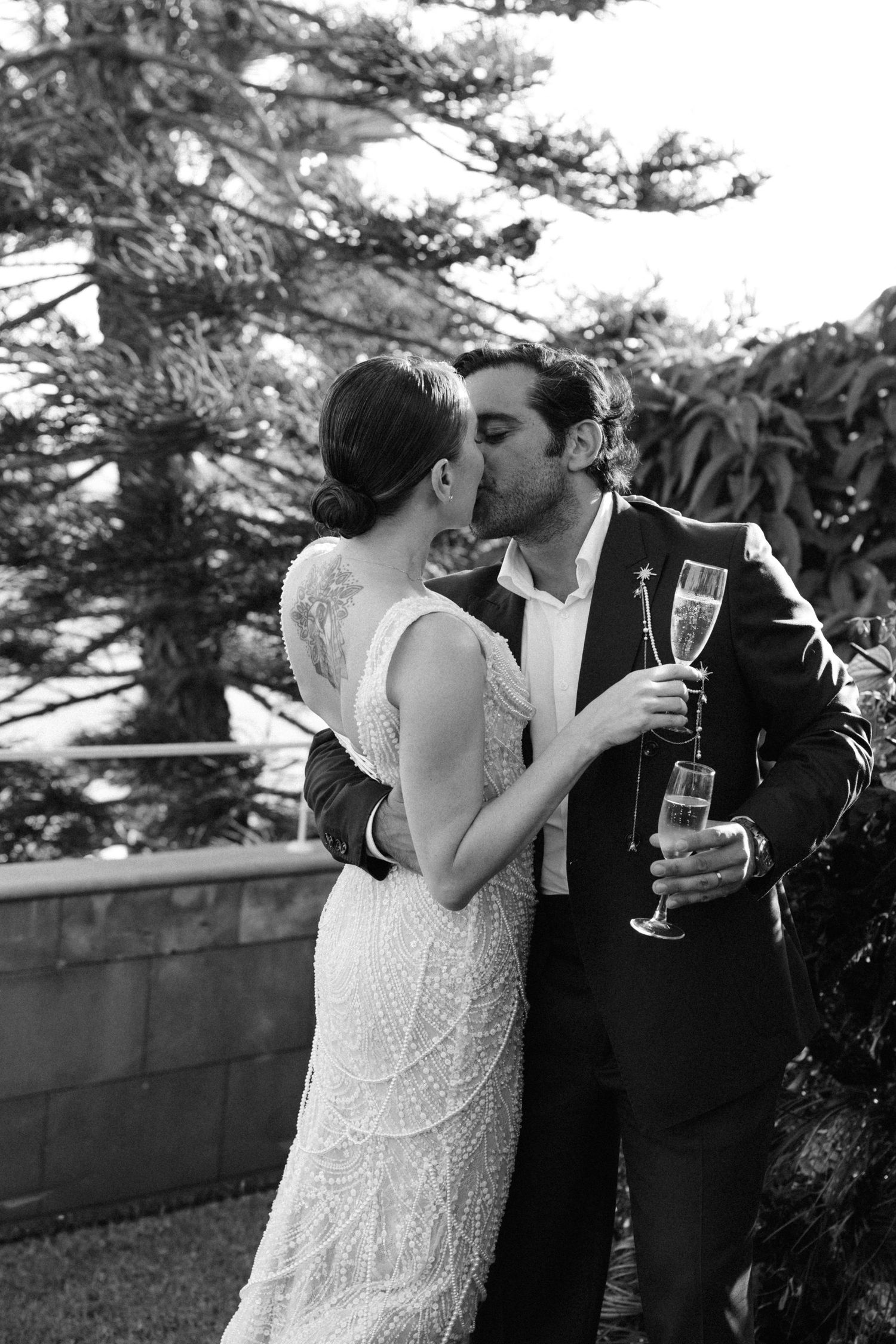 Bride and groom share a romantic kiss while holding champagne glasses outdoors in black and white wedding photo.