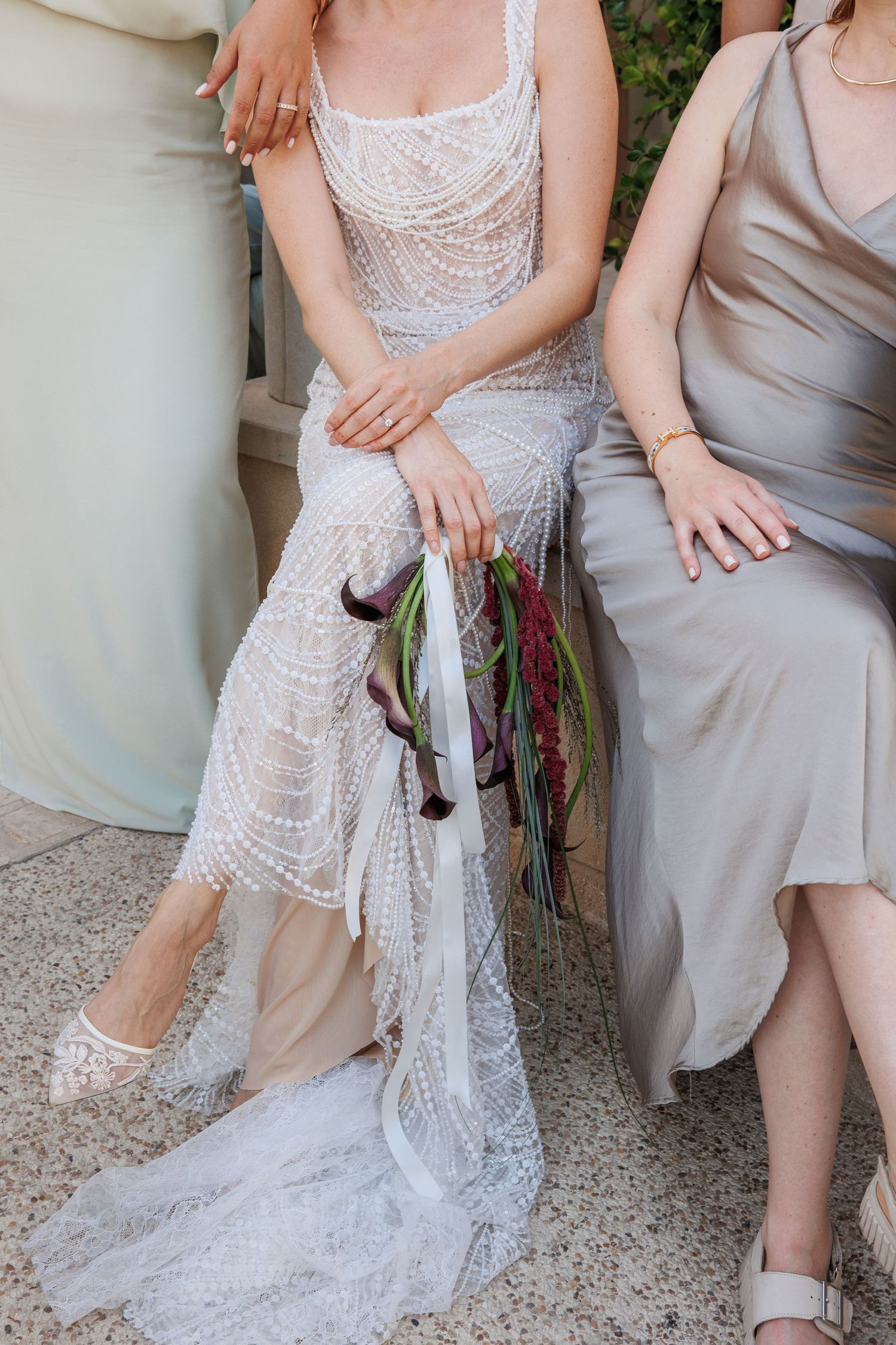 Bride in beaded lace wedding gown holding cascading floral bouquet, seated beside bridesmaid in silver dress.
