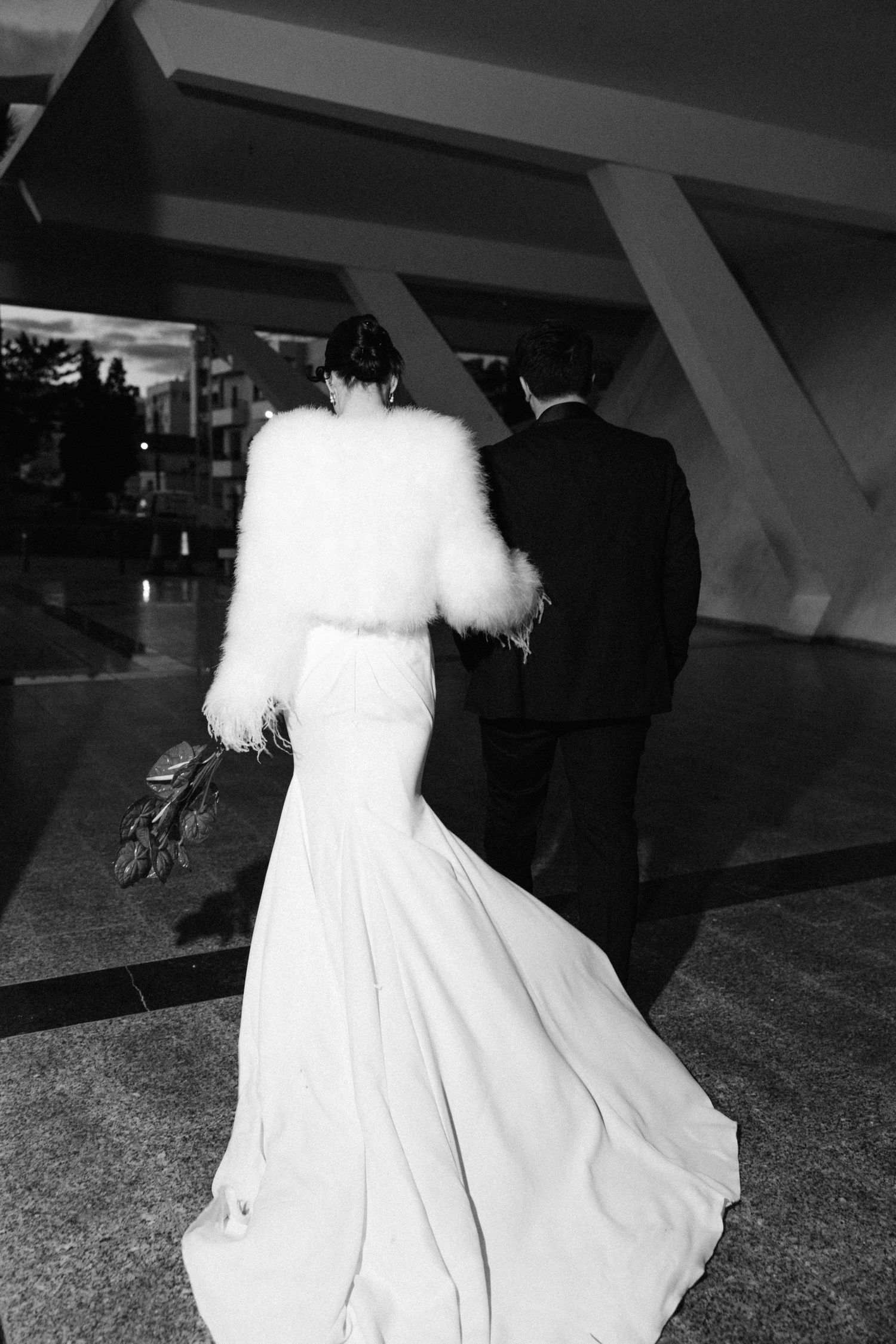 Black and white photo of bride in white gown and feather jacket walking with groom into a modern venue.