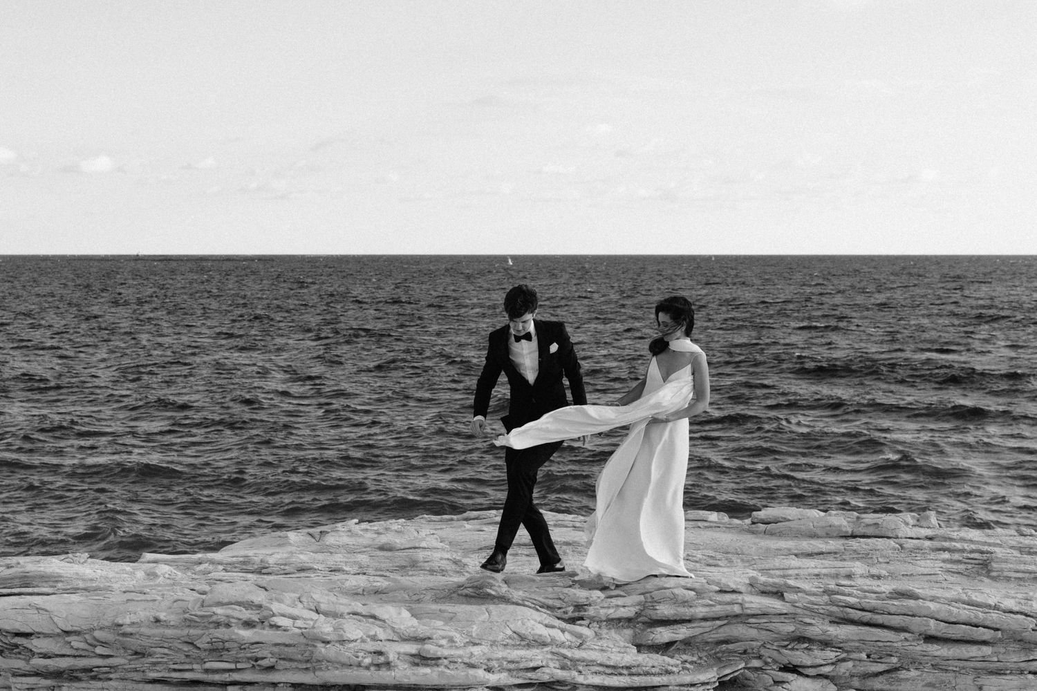 A couple walks together along the beach shore in black and white as ocean waves roll in.