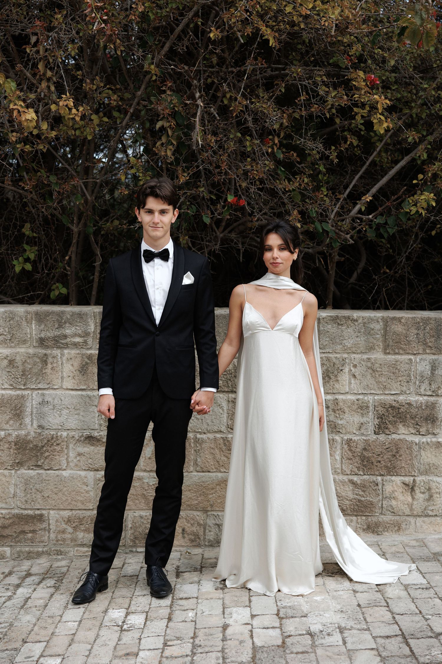 Elegant couple in formal wedding attire standing together against stone wall and trees outdoors.