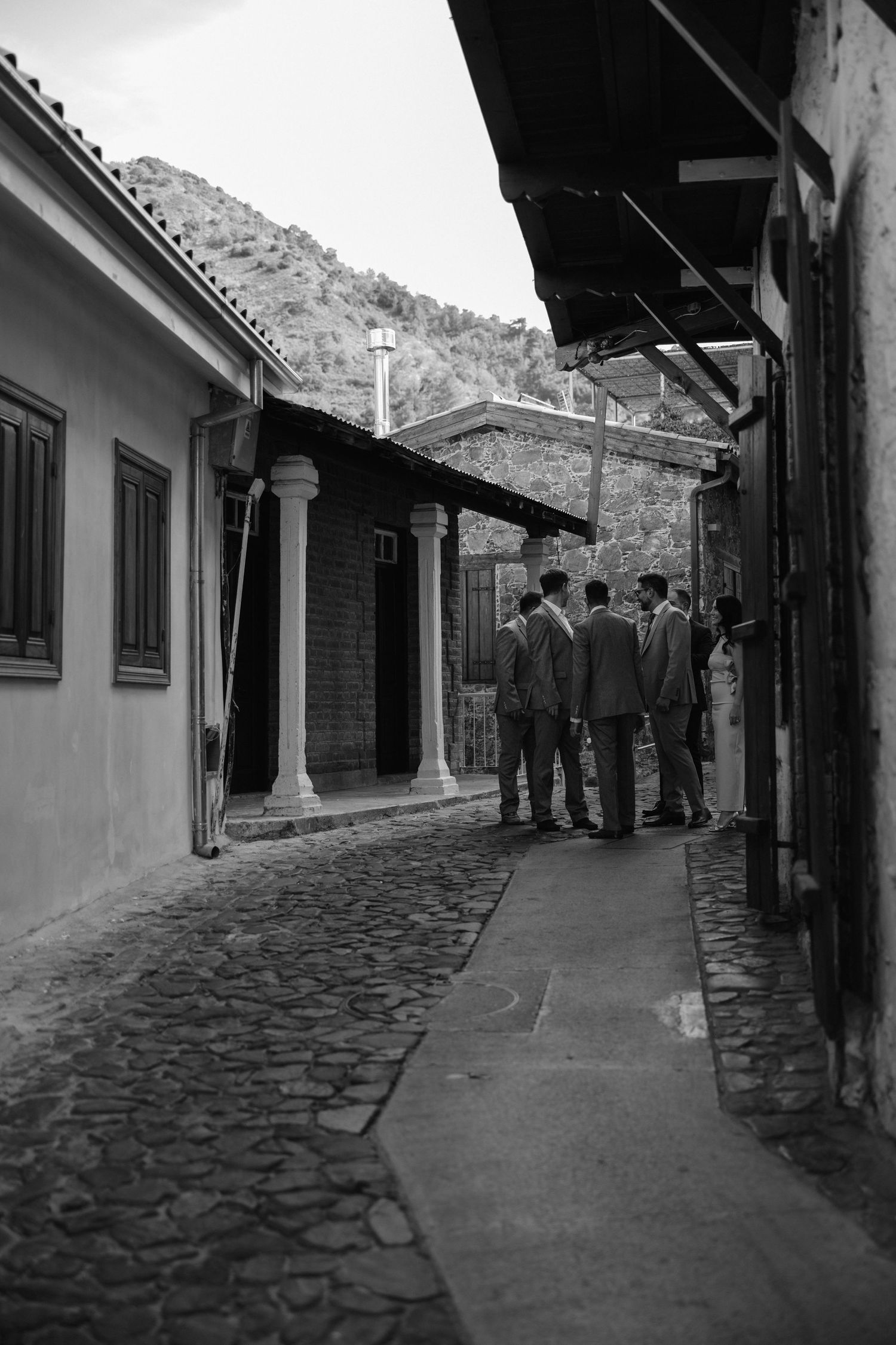 A group of people gather in a narrow cobblestone alley between old buildings with a mountain visible in the background.