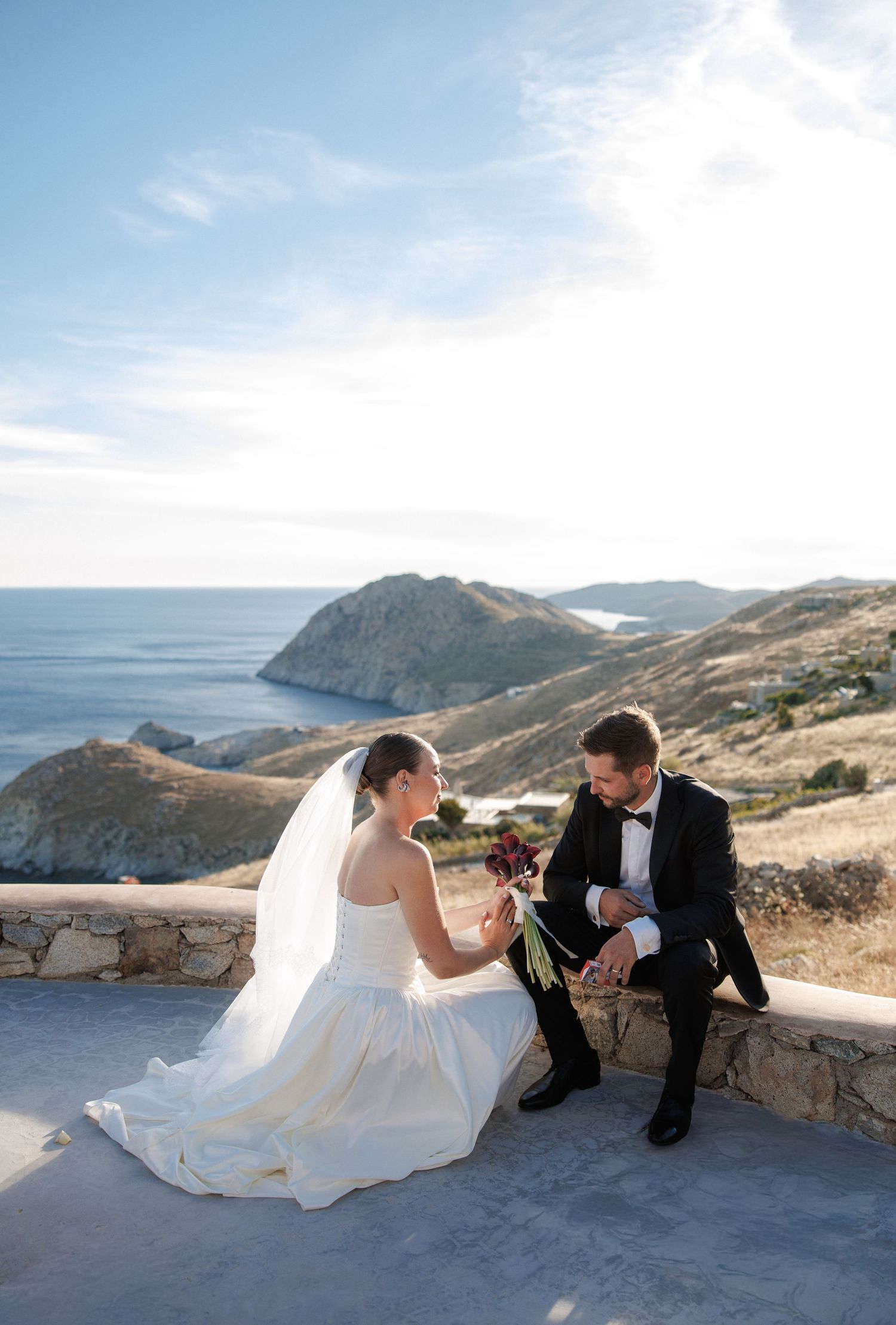 Bride and groom sit together on a stone terrace overlooking a scenic coastal landscape on their wedding day.