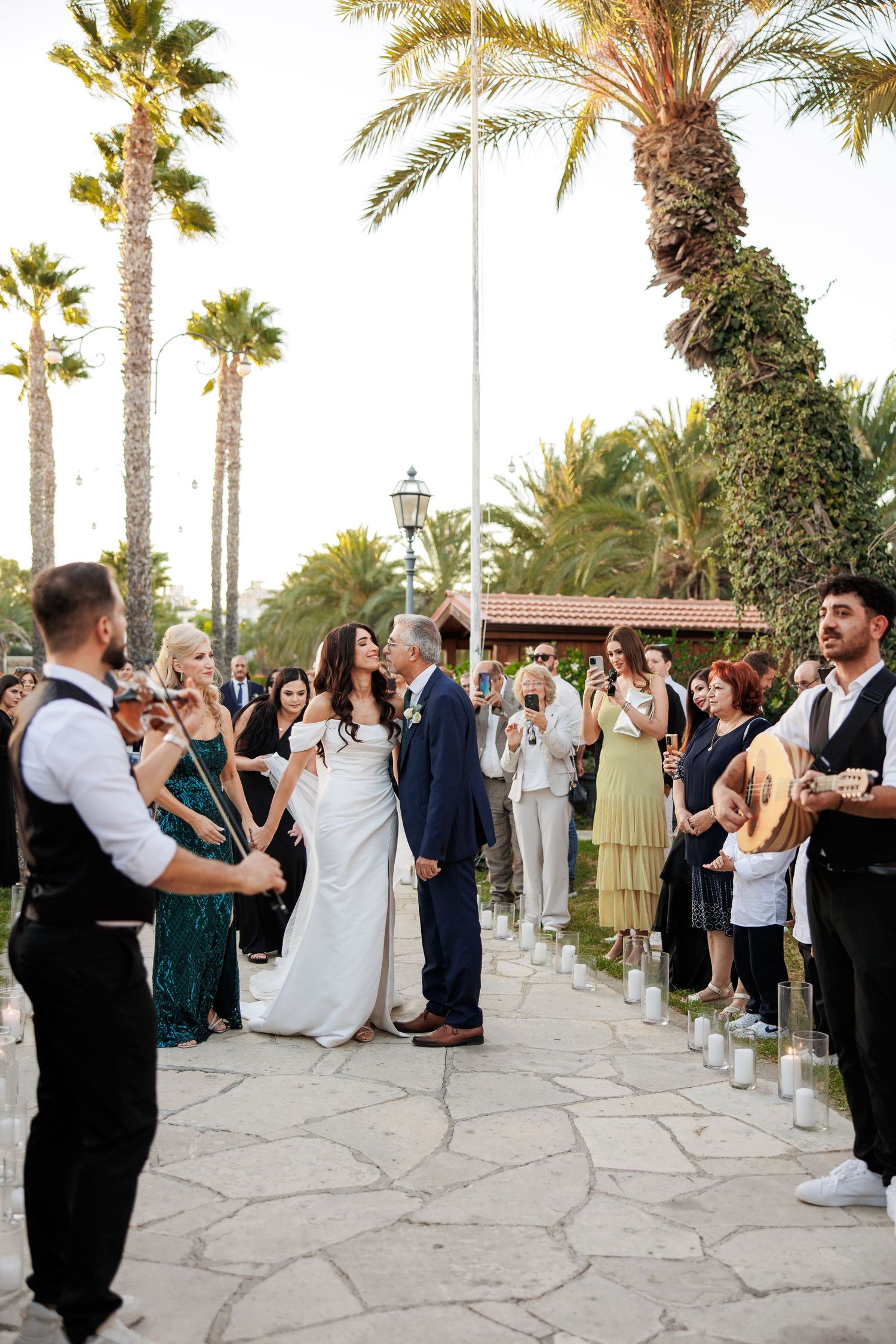 Bride and groom share a first dance outdoors surrounded by guests and palm trees at a Mediterranean wedding.