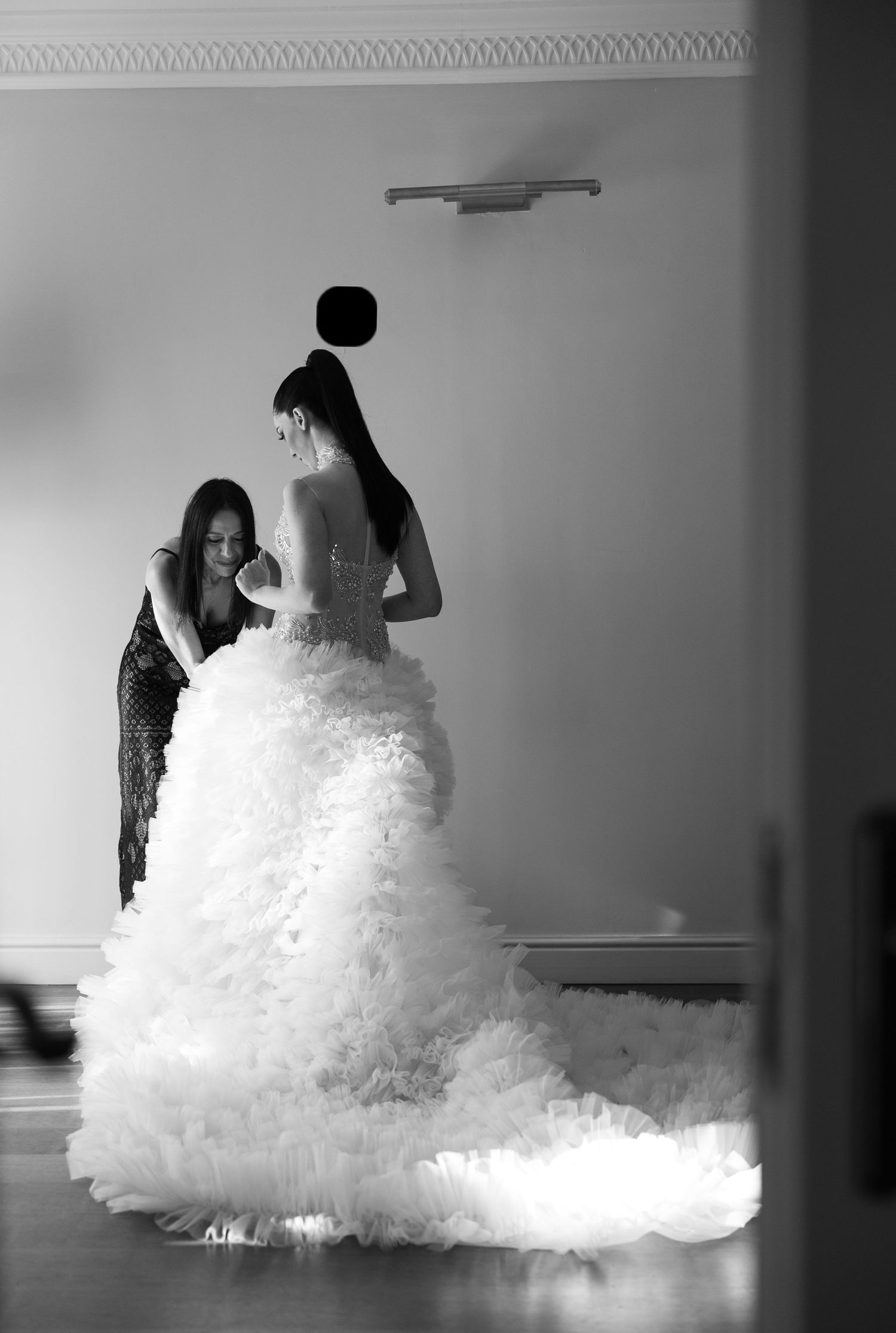 Black and white photo of a bride in a ruffled wedding gown being assisted by a bridesmaid in an elegant room.