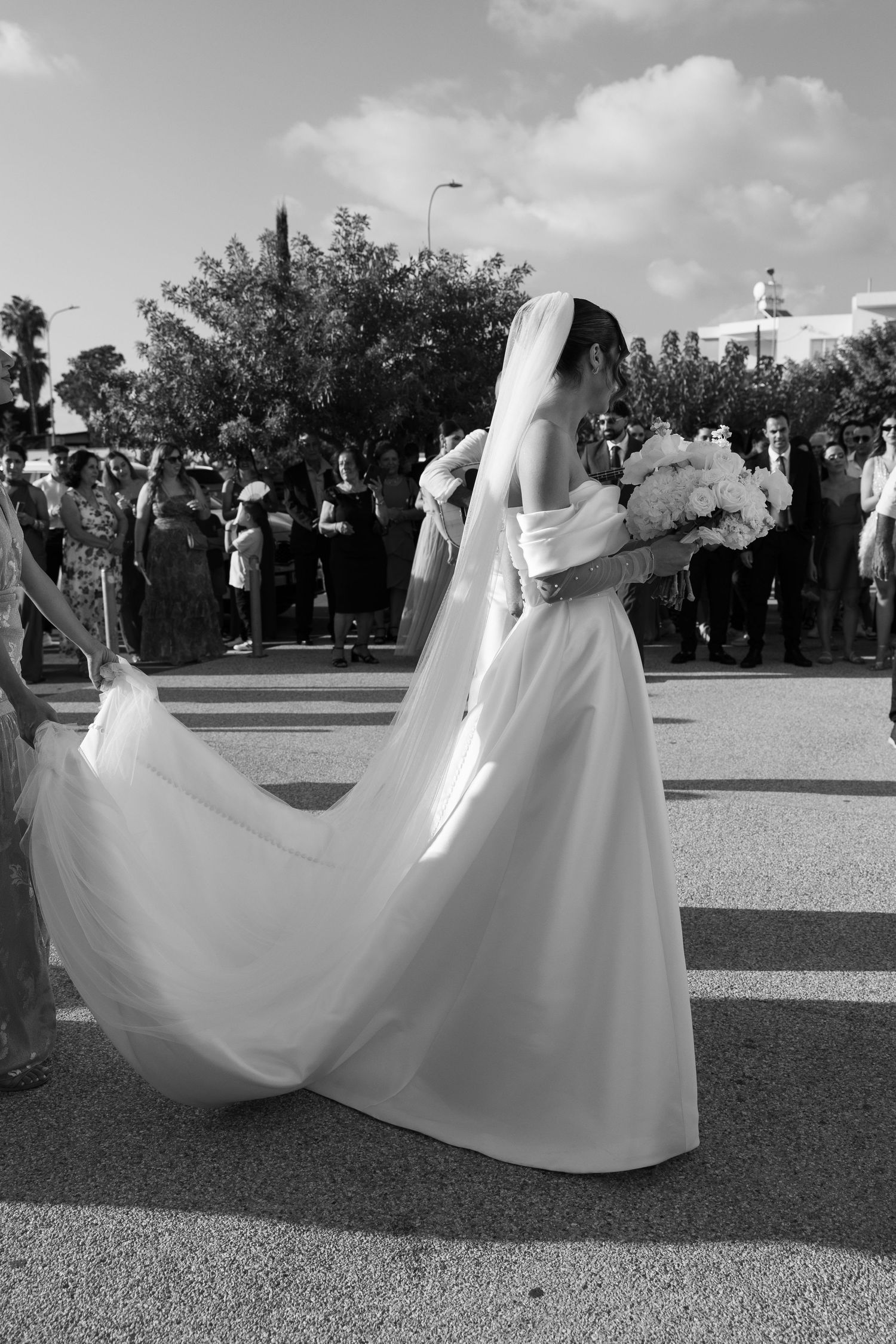 Bride in white gown and veil holds bouquet while guests gather in background, black and white wedding photo.