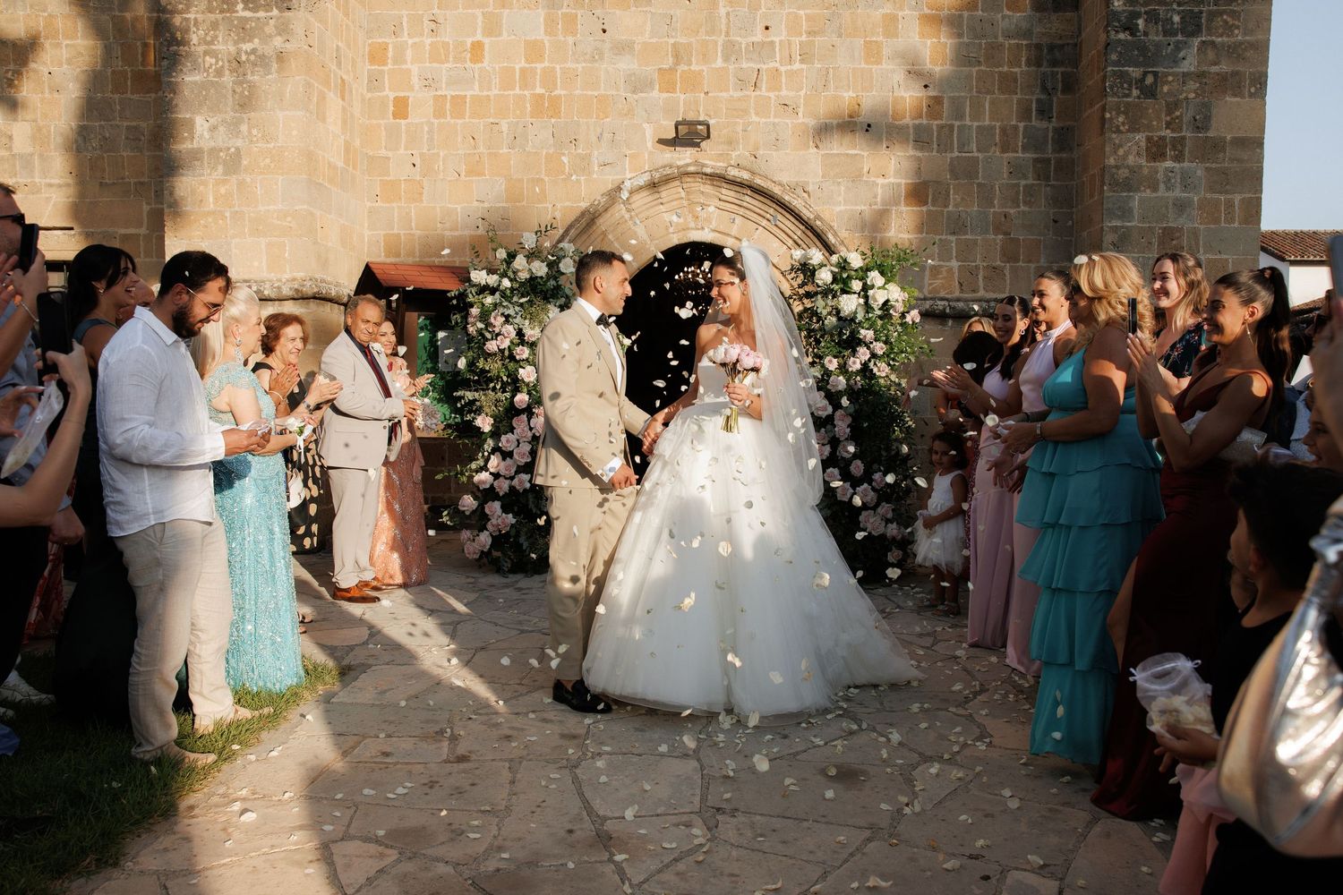 Wedding guests celebrate under a stone archway decorated with white floral arrangements.