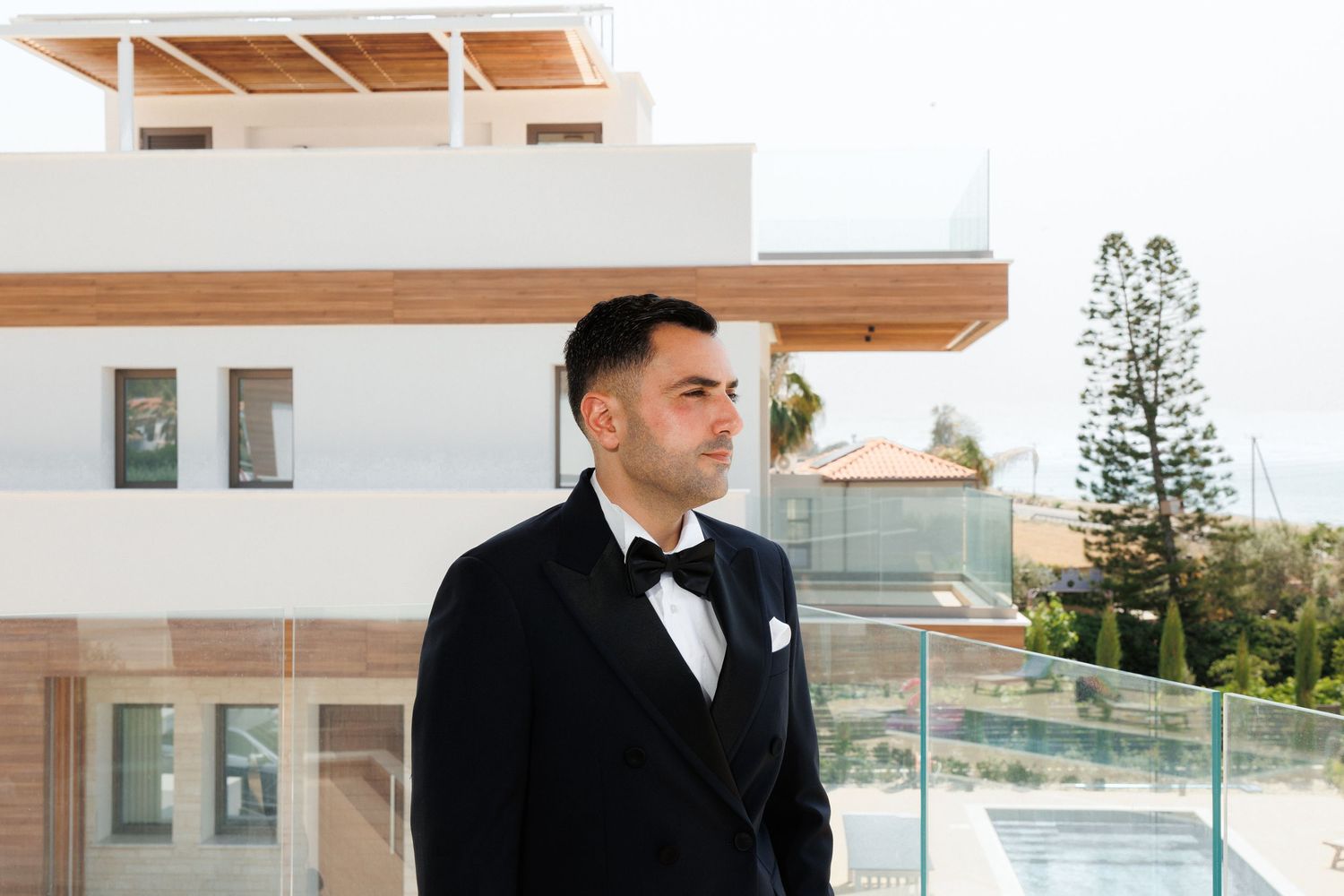 A person in a black tuxedo with bowtie stands on a balcony of a modern building during daytime.