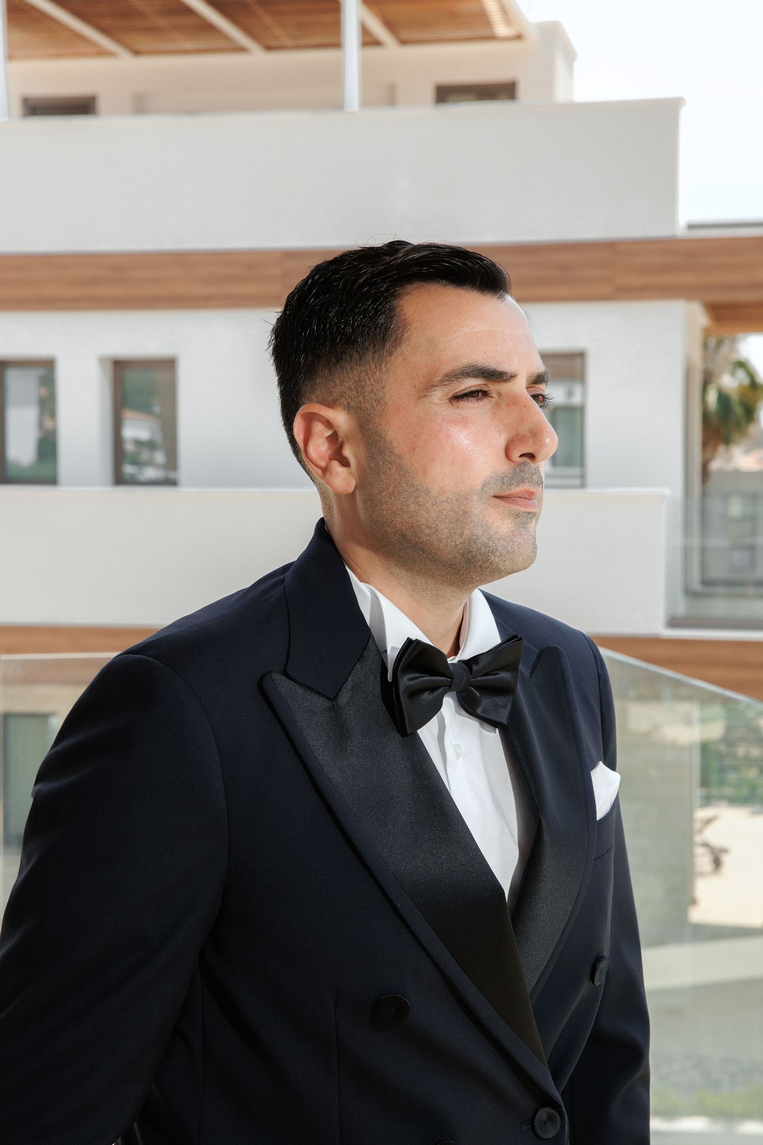 Profile view of a groom in a classic black tuxedo with bow tie and white pocket square looking off into the distance.