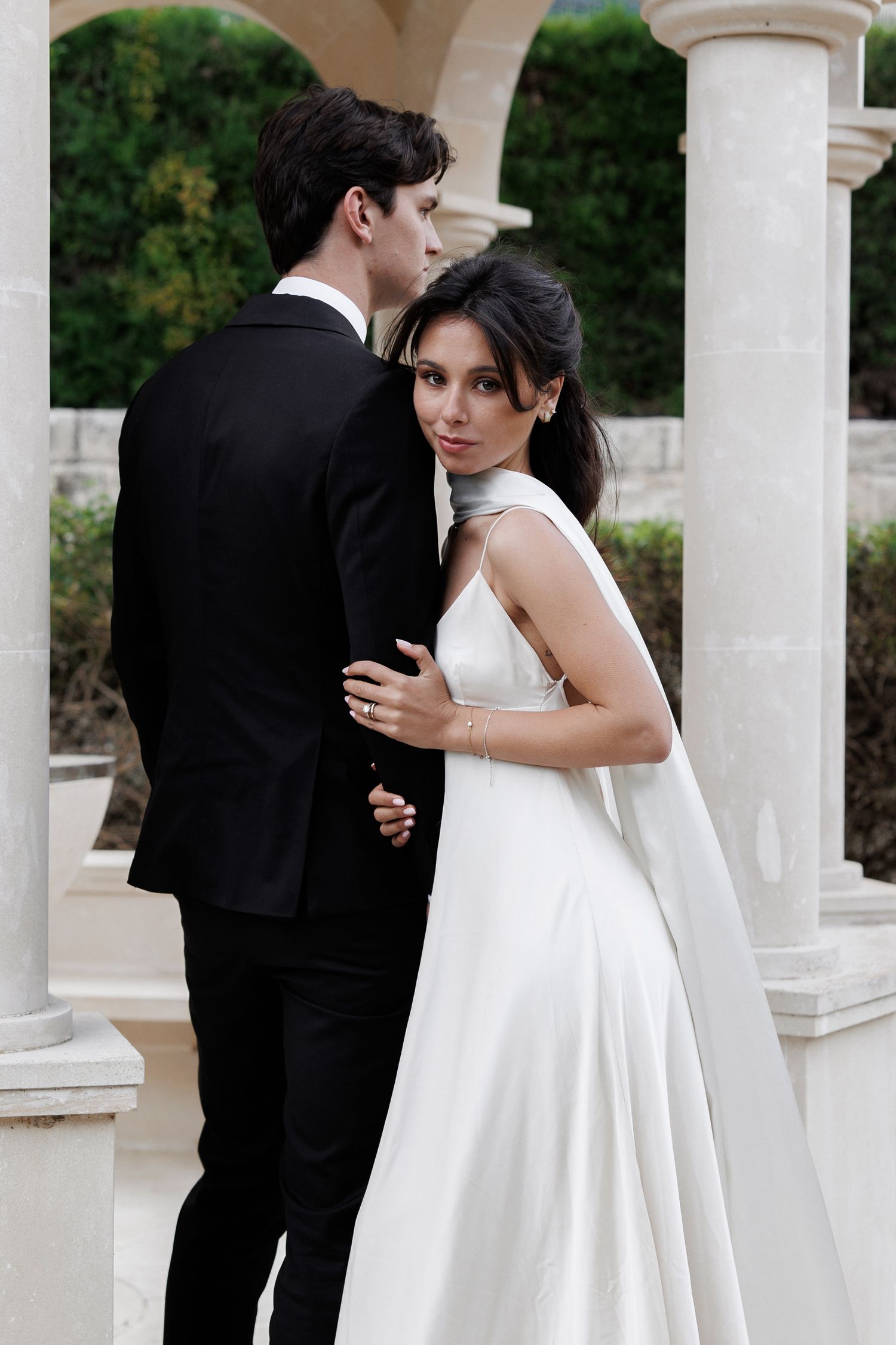 A wedding couple embraces under white columns in an elegant outdoor setting with the bride in a flowing white gown.
