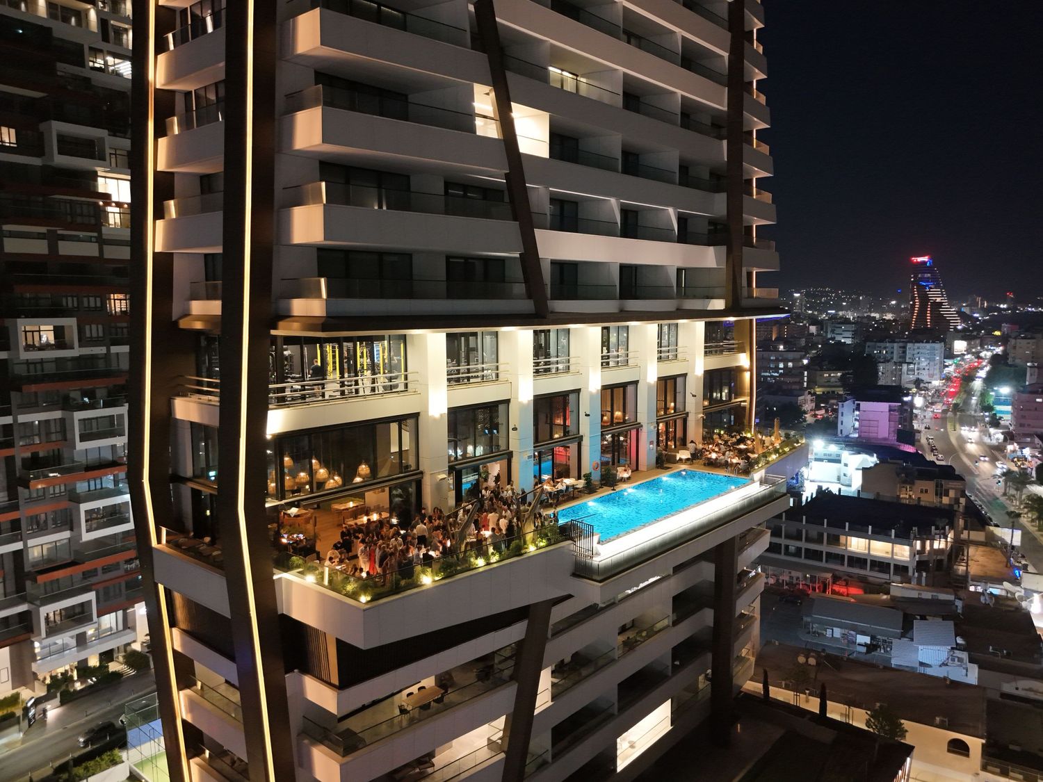 A high-rise building at night with an illuminated rooftop pool and lounge area overlooking a cityscape.