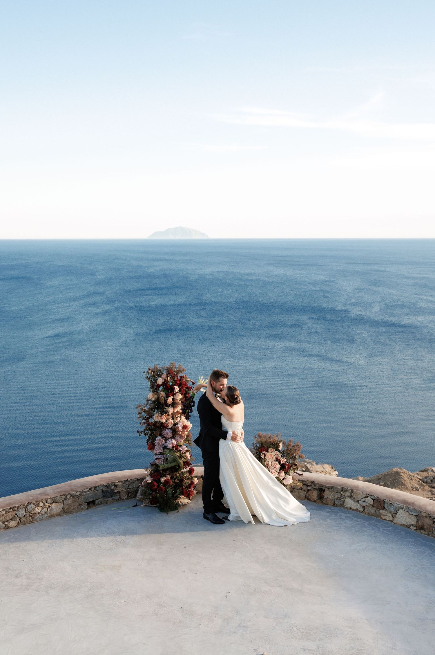 Couple embracing at seaside wedding ceremony with floral arch overlooking blue ocean horizon.