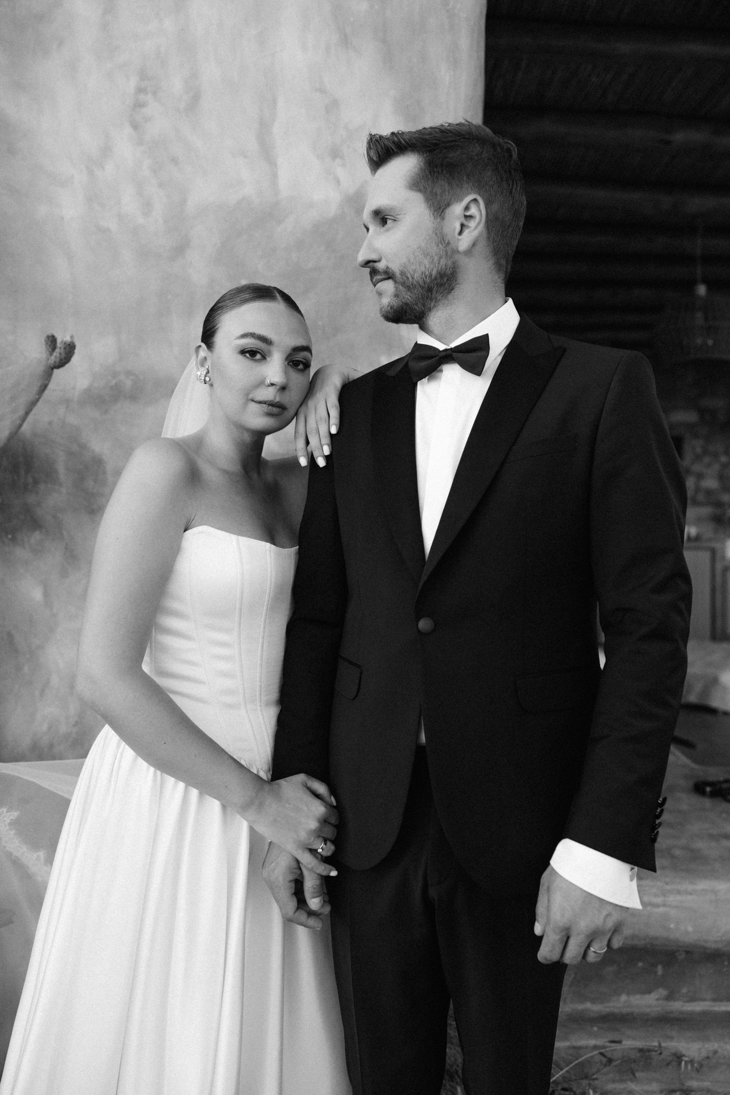 Elegant bride and groom pose in black and white portrait, she in strapless gown, he in classic black tuxedo with bow tie.