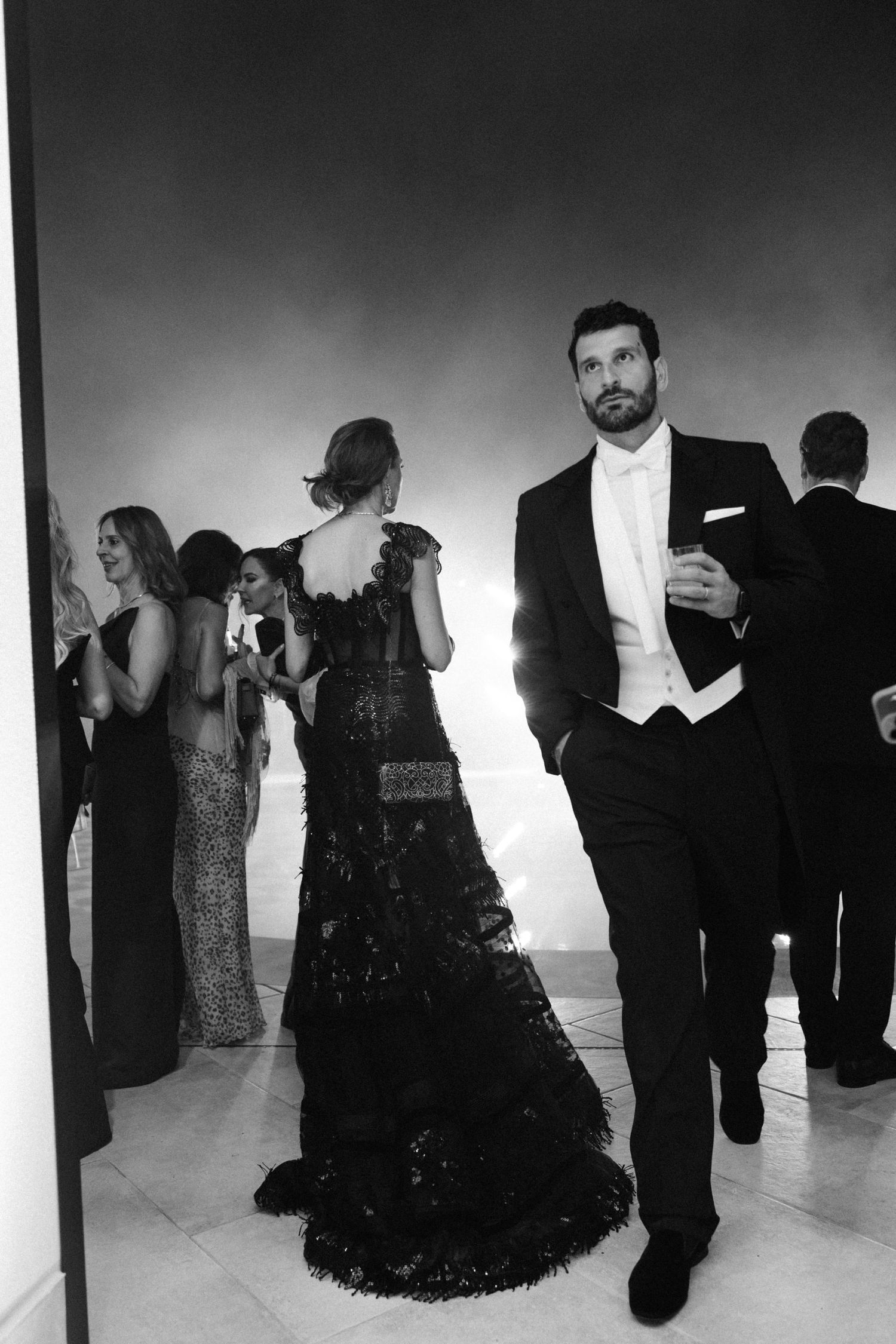 Black and white photo of elegant guests at a formal gala, man in tuxedo walks forward as women in gowns gather behind.