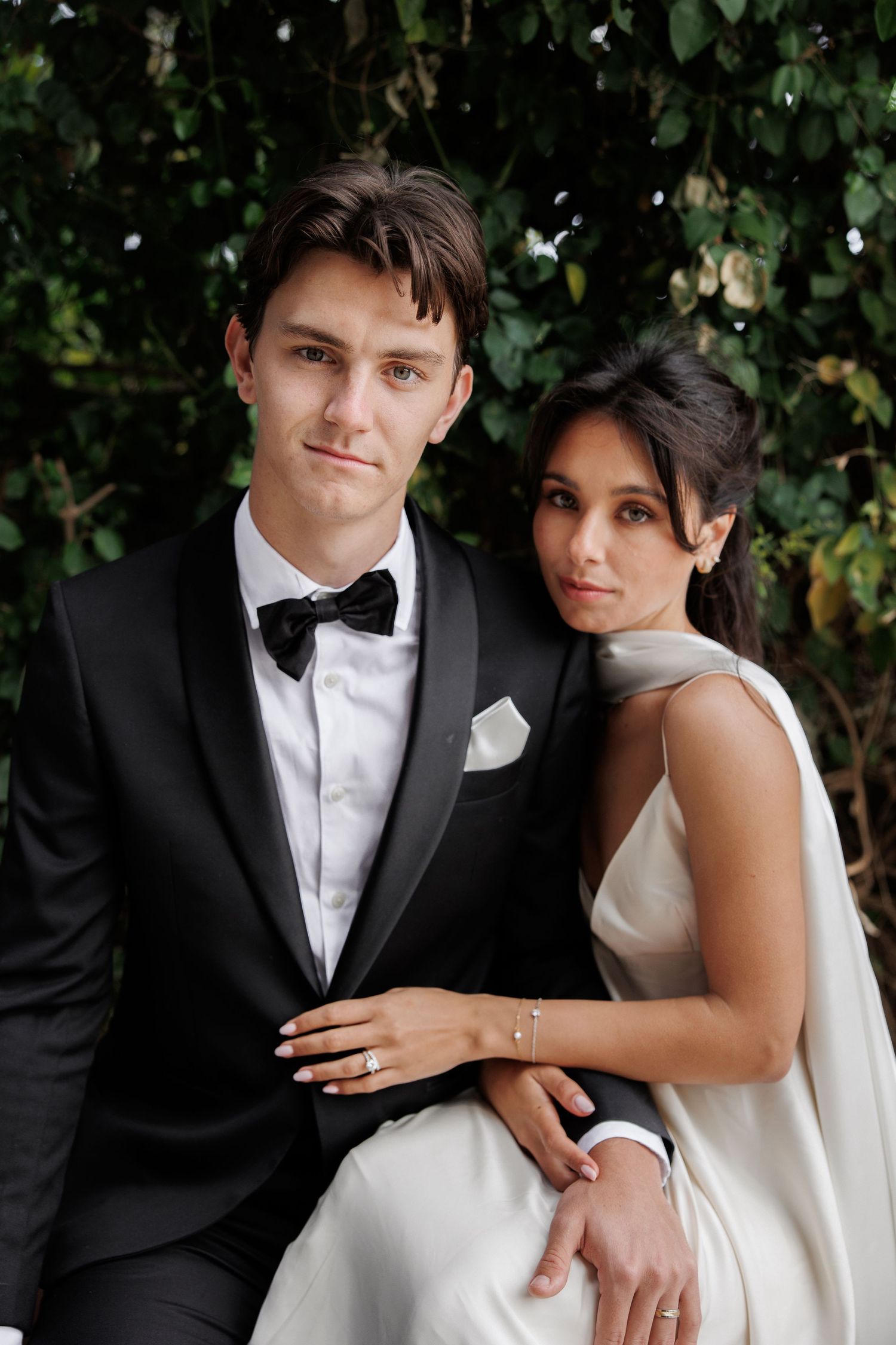 A formal wedding photo of a couple in black tuxedo and white dress posed against a green foliage background.