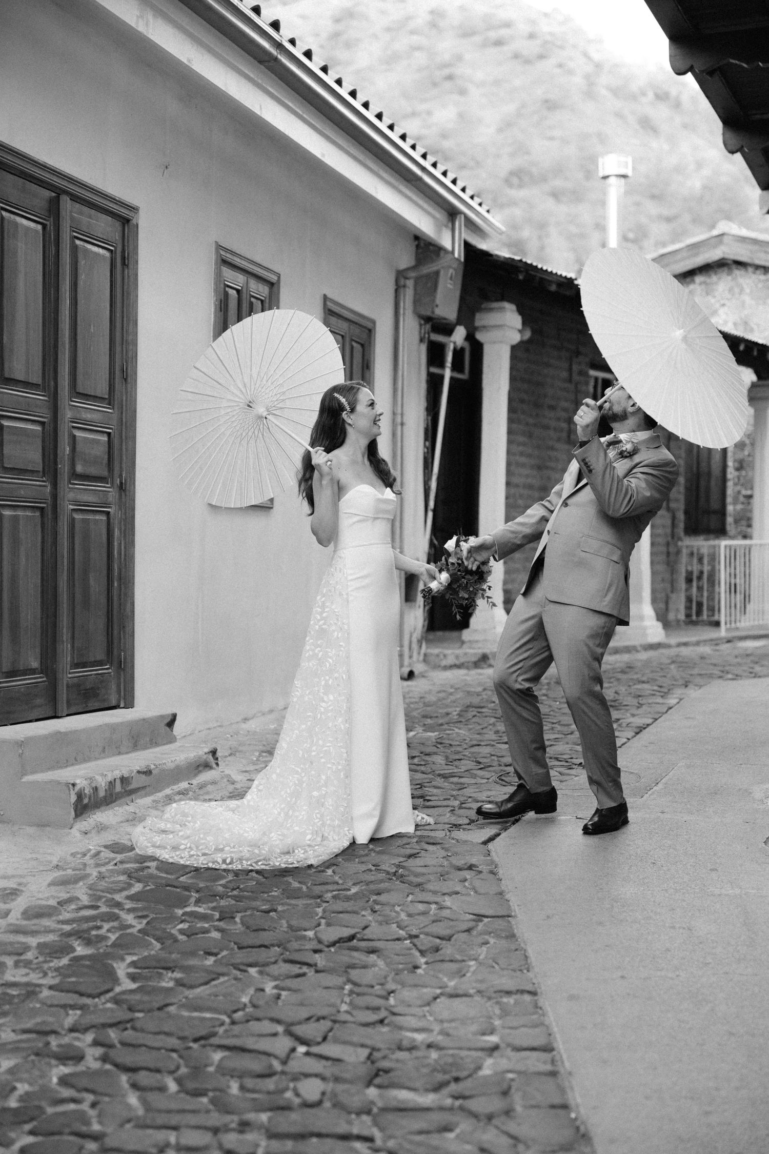 Black and white photo of couple with parasols sharing playful moment on cobblestone street.