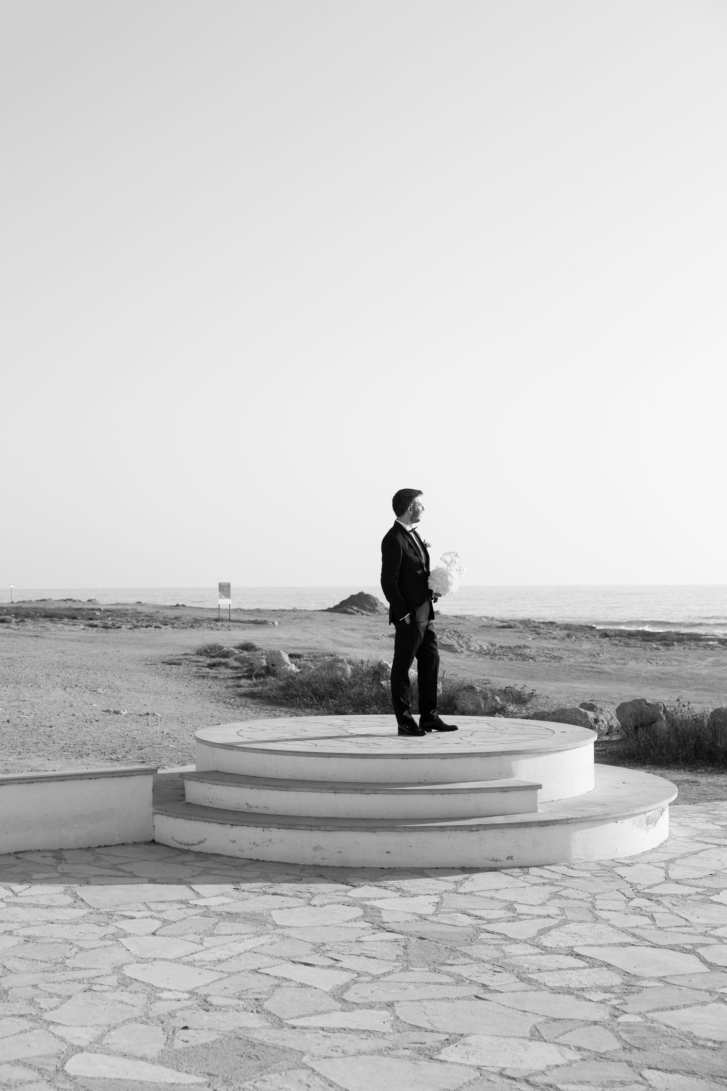 Man in black suit standing on circular white platform near beach in black and white photo.