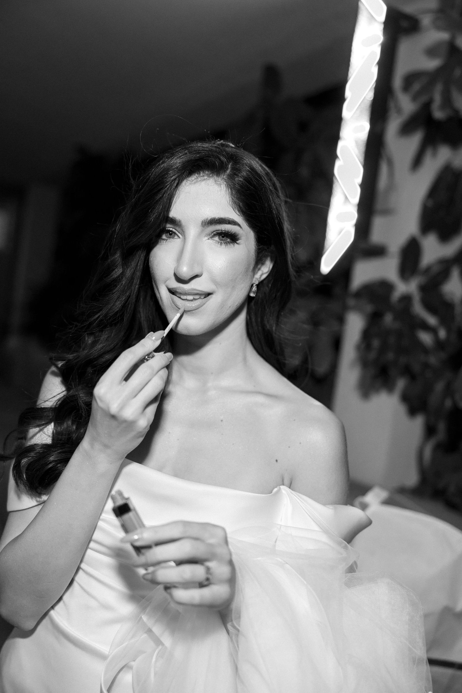 Black and white photo of a bride applying lipstick while wearing an off-shoulder wedding dress under bright studio lighting.