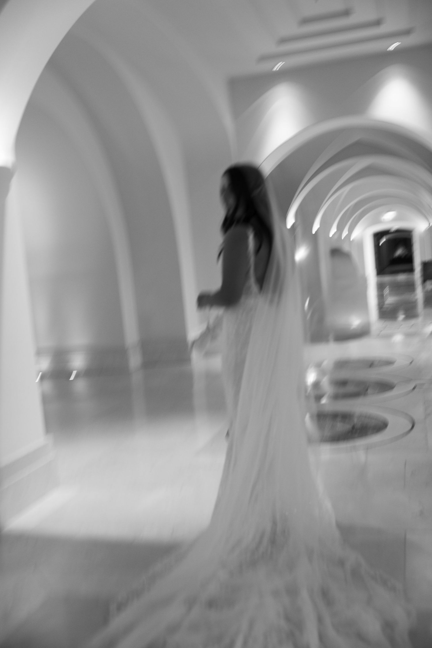 Blurred black and white photo of bride in flowing gown walking through arched hotel corridor.