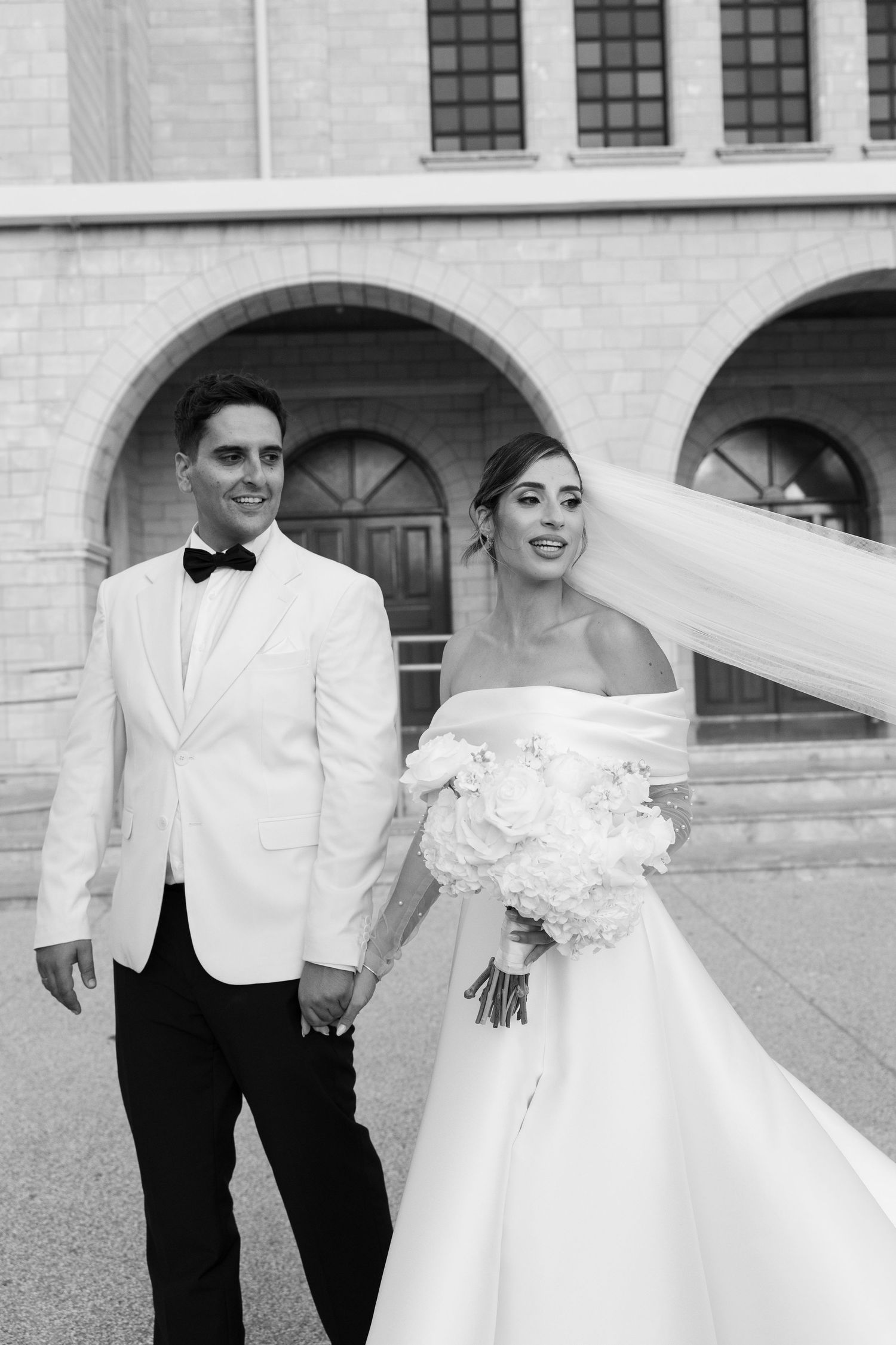Black and white photo of elegant bride and groom holding hands outside a grand building with arched windows.