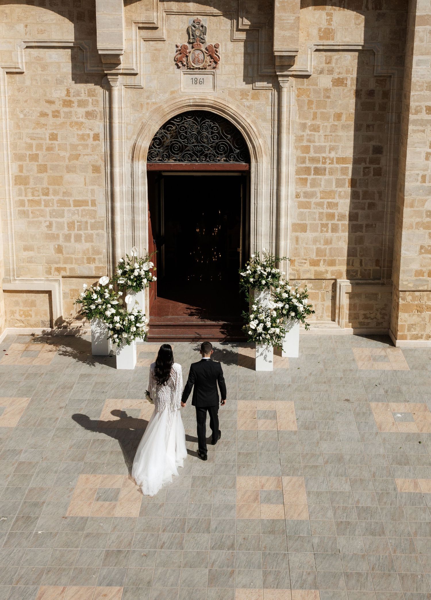 Bride and groom walking hand in hand toward an ornate church entrance adorned with white floral arrangements.