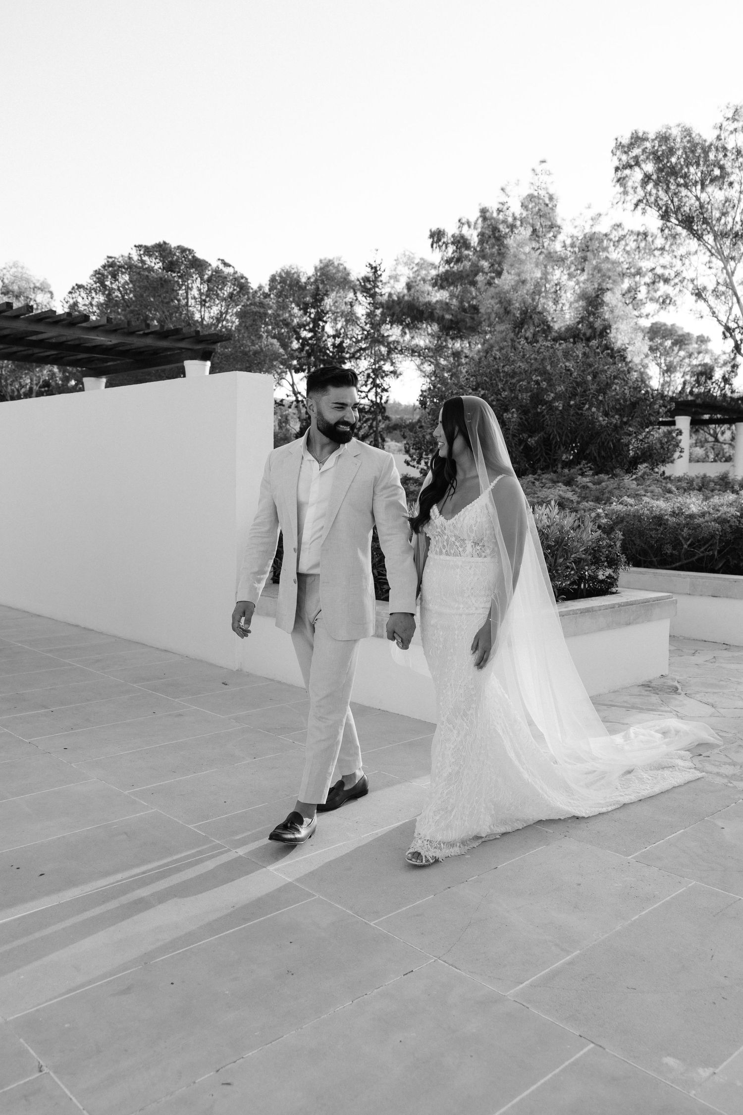 Black and white photo of bride and groom walking hand in hand on a terrace at their outdoor wedding.
