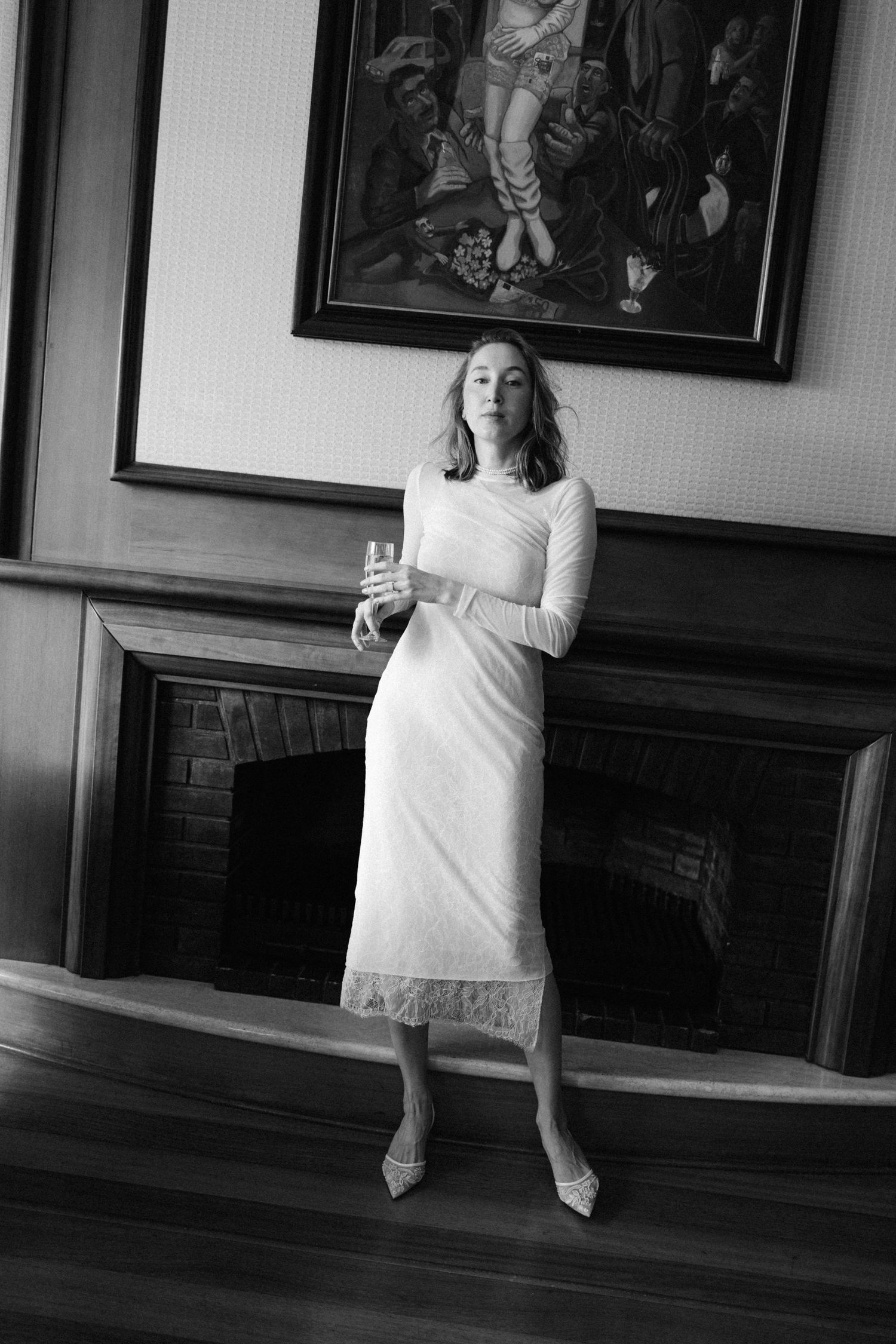 Woman in white lace midi dress holding champagne glass by fireplace, black and white portrait photo.
