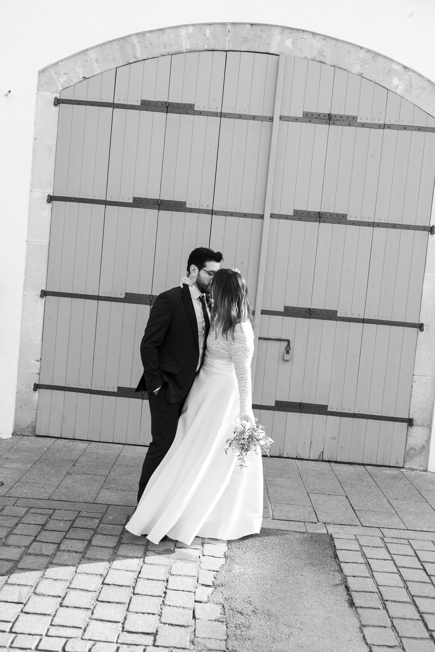 Black and white wedding photo of bride and groom kissing before large arched wooden doors on cobblestone.