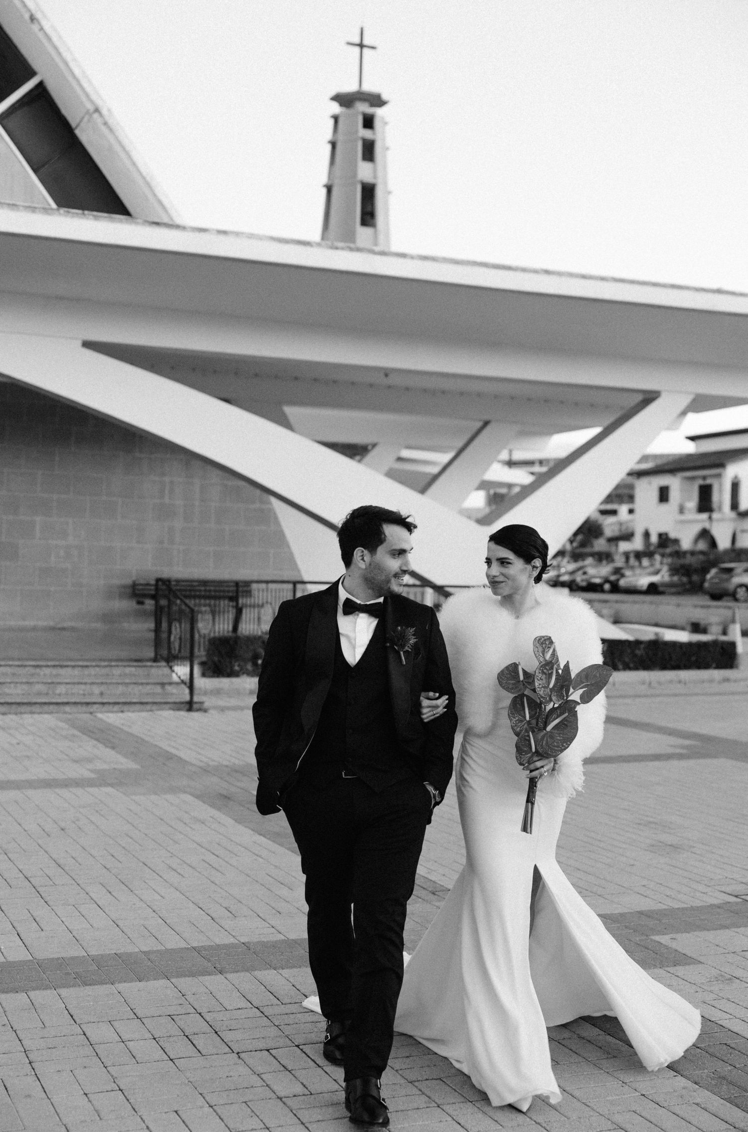 Black and white photo of bride and groom walking together outside a church, bride holding bouquet in elegant white gown.