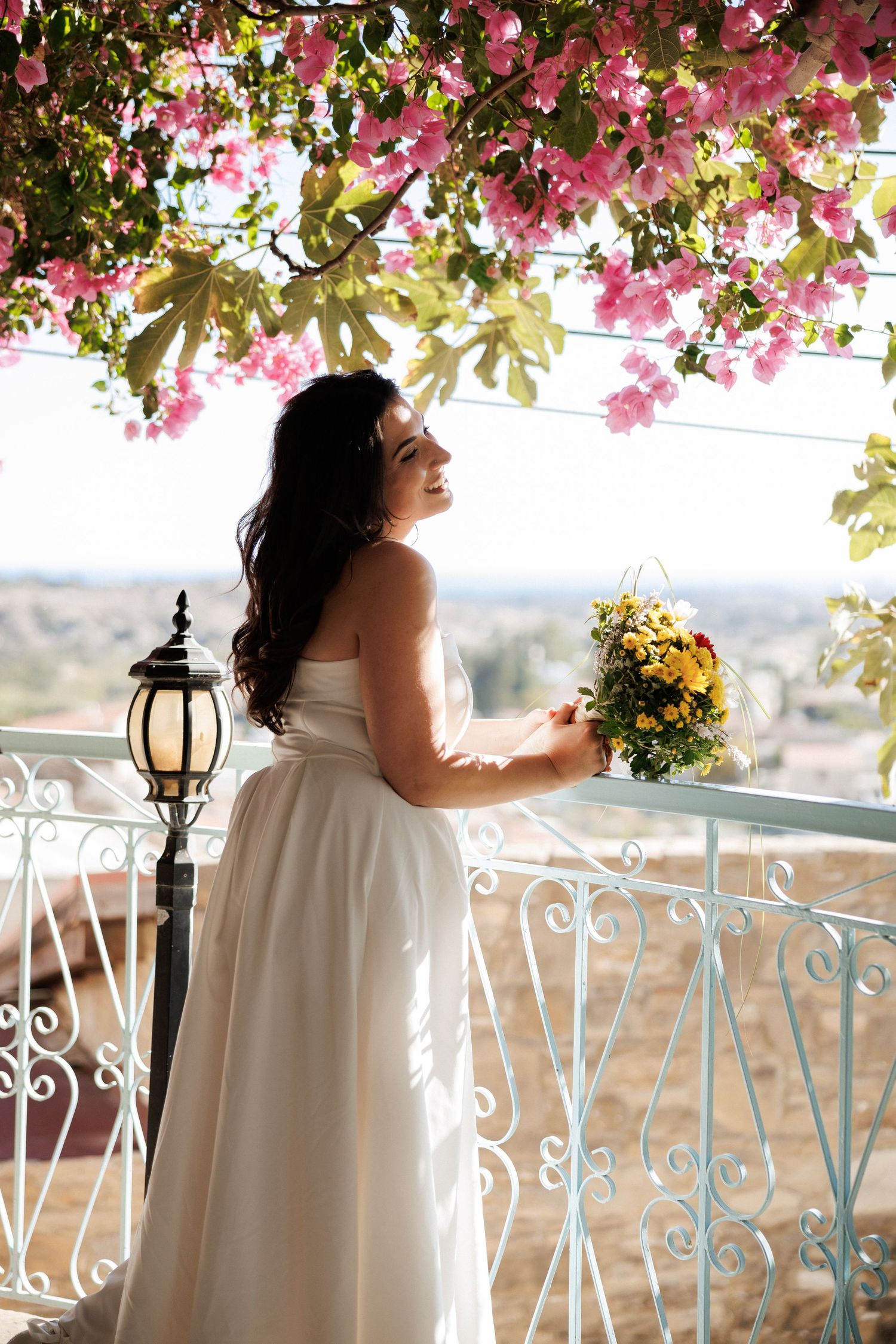 A bride holding a bouquet stands on a balcony under pink bougainvillea flowers.