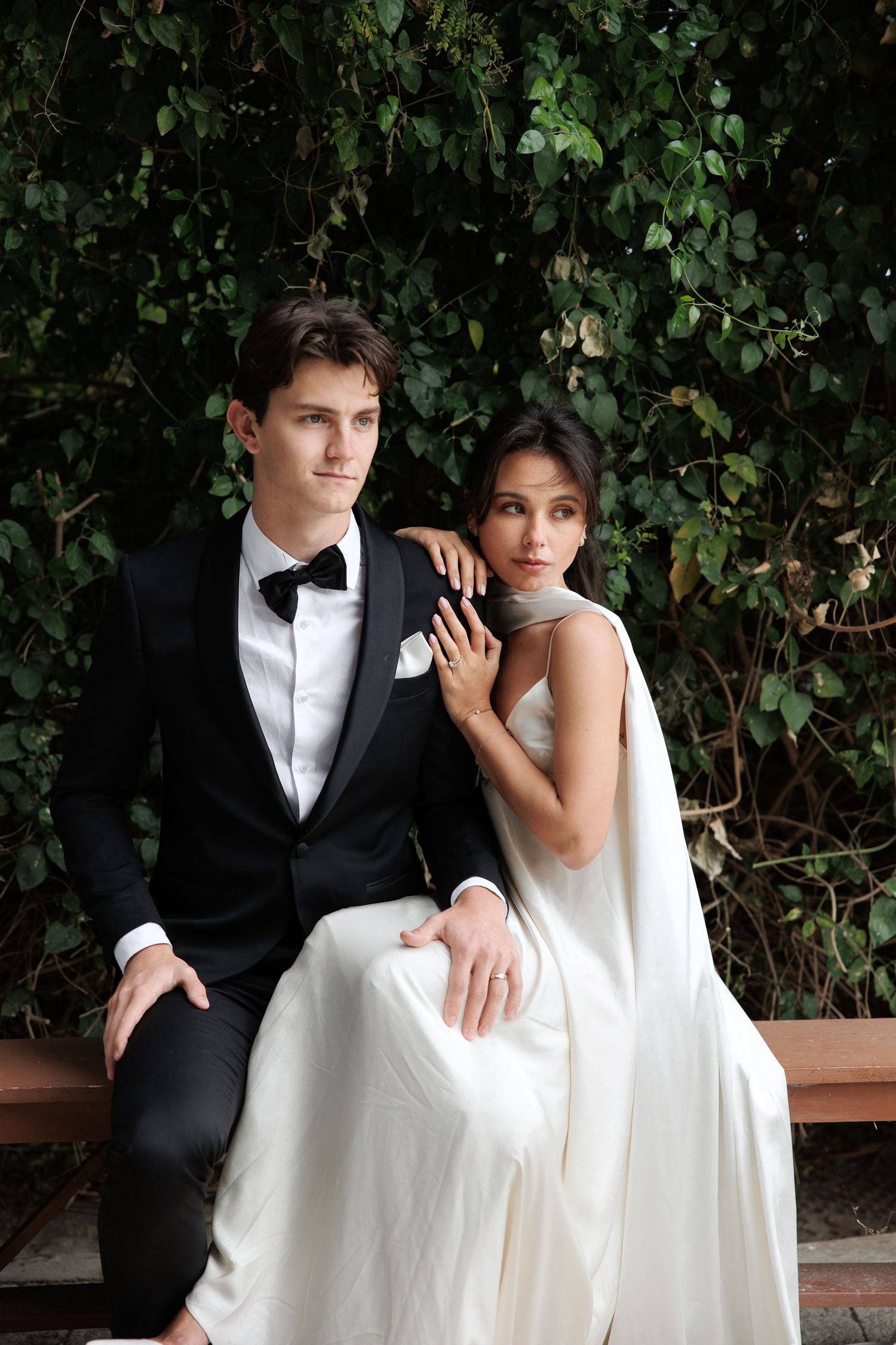 Elegant couple sits on bench against ivy wall, bride in white dress and groom in classic tuxedo.