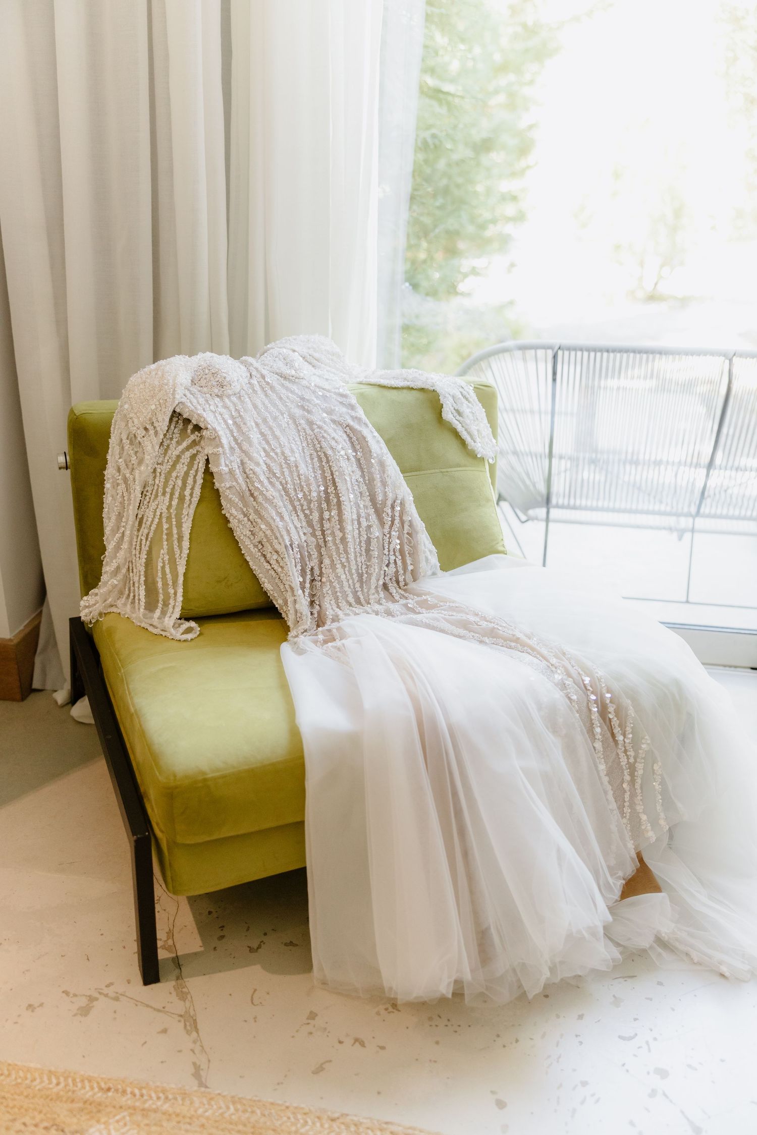 A white beaded wedding dress draped over a green velvet chair near a bright window.