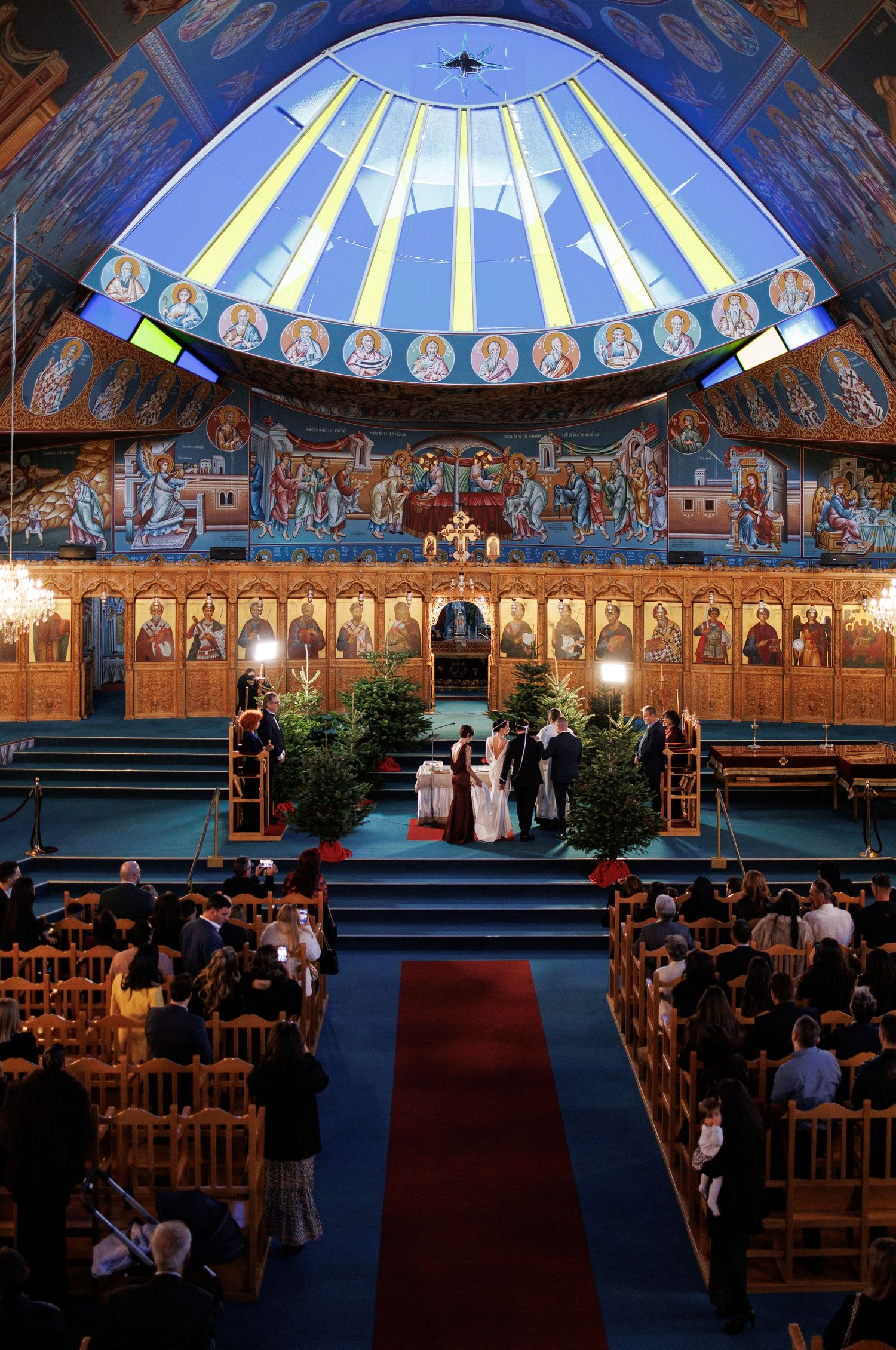 Orthodox church wedding ceremony with ornate Byzantine frescoes, blue dome, and red carpet aisle filled with guests.