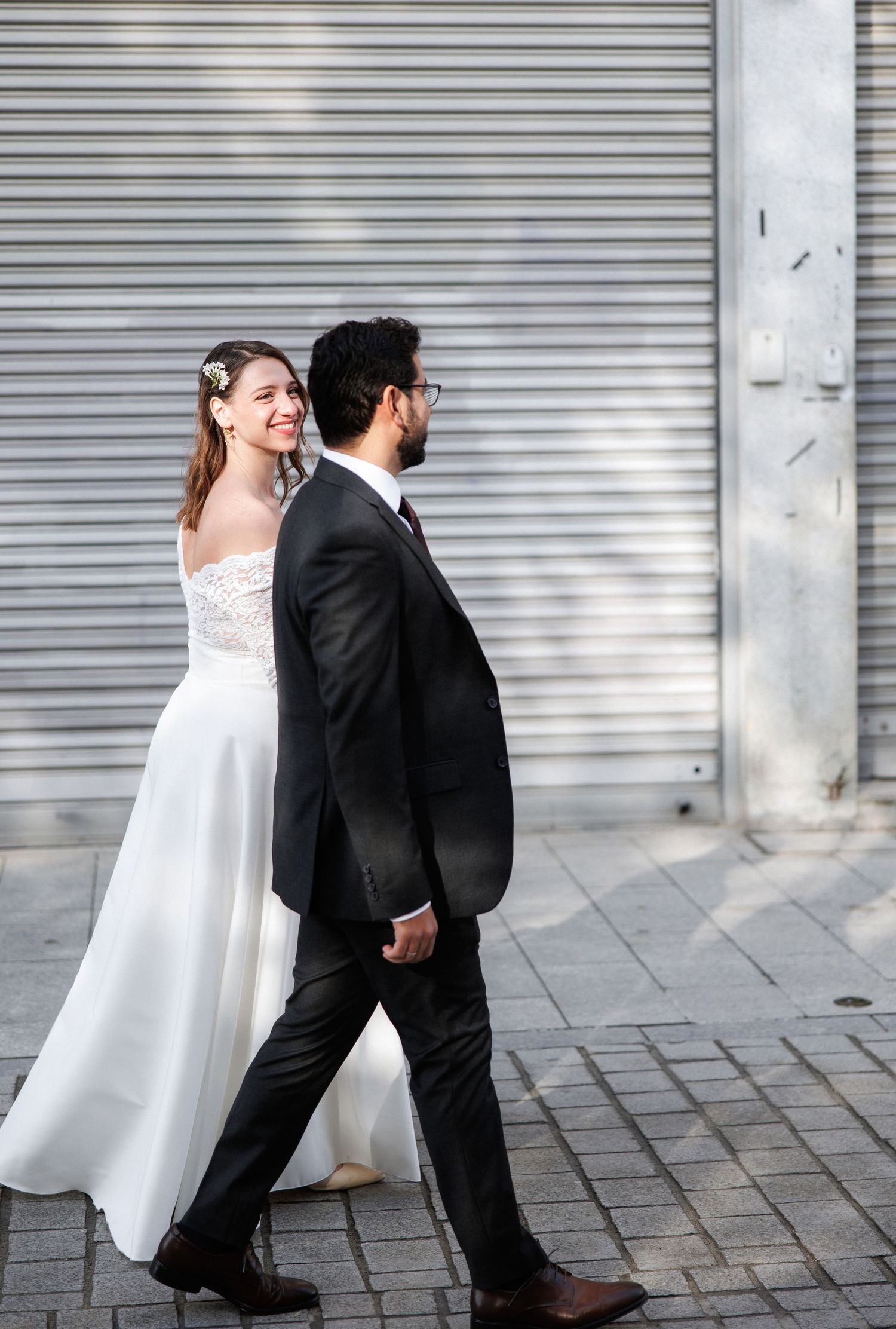 Smiling bride in white gown holds hands with groom in black suit walking on urban street.