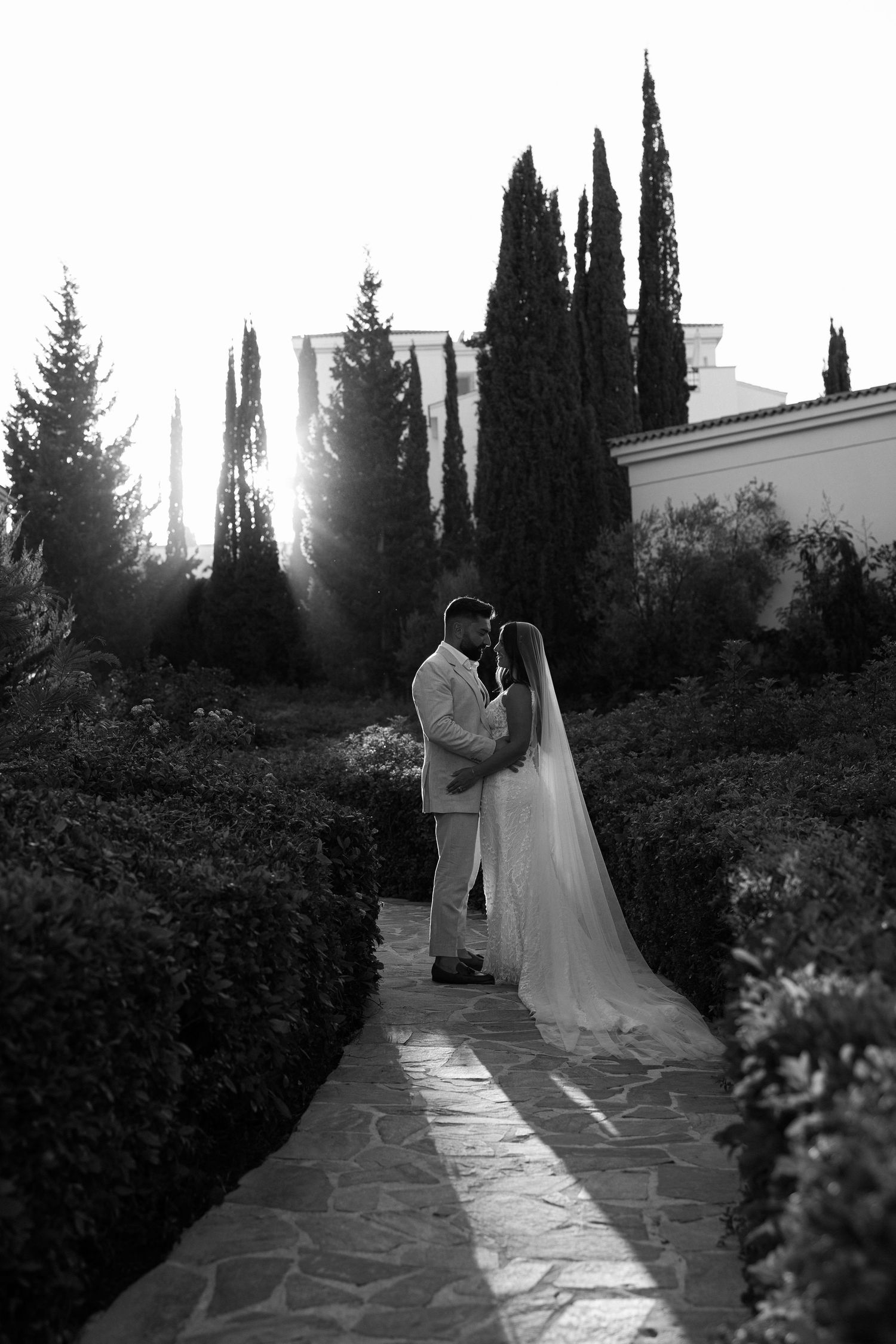 Black and white photo of bride and groom embracing on garden path with cypress trees and sunlight behind them.
