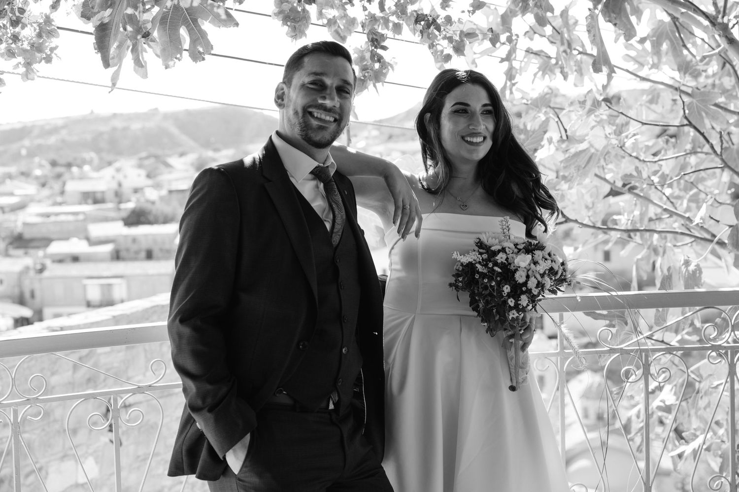 A black and white wedding photo of a couple posing on a balcony overlooking a scenic view.