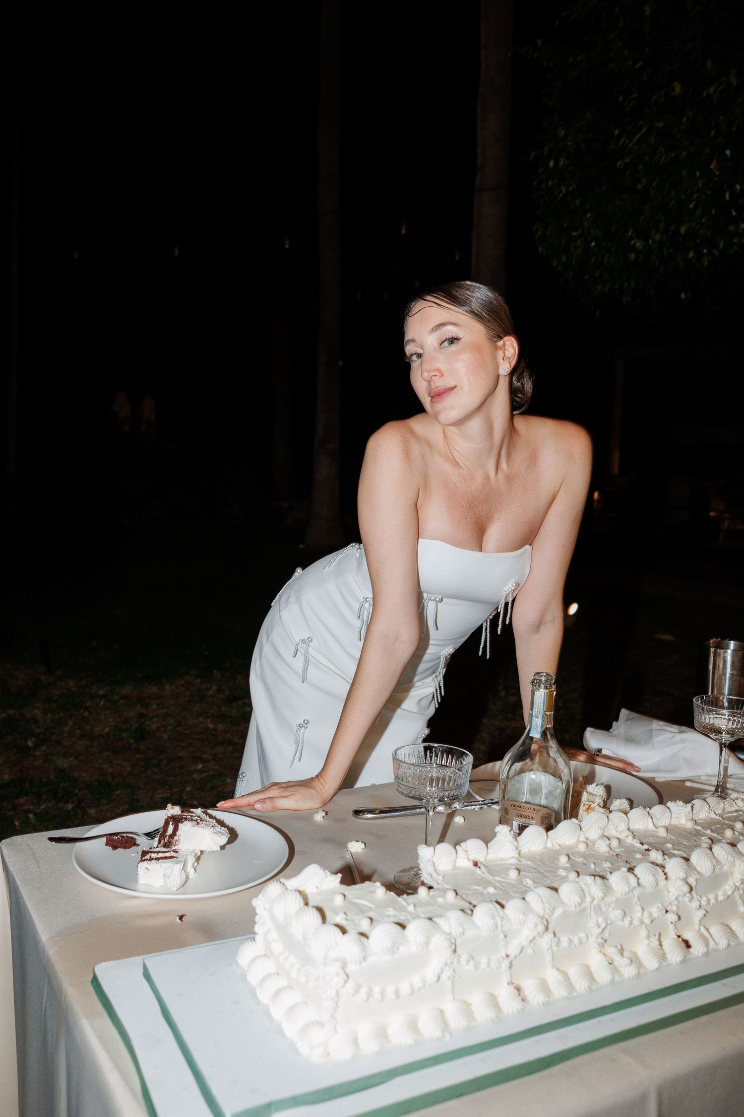 Bride in strapless white gown leaning over table with decorated wedding cake at night reception.