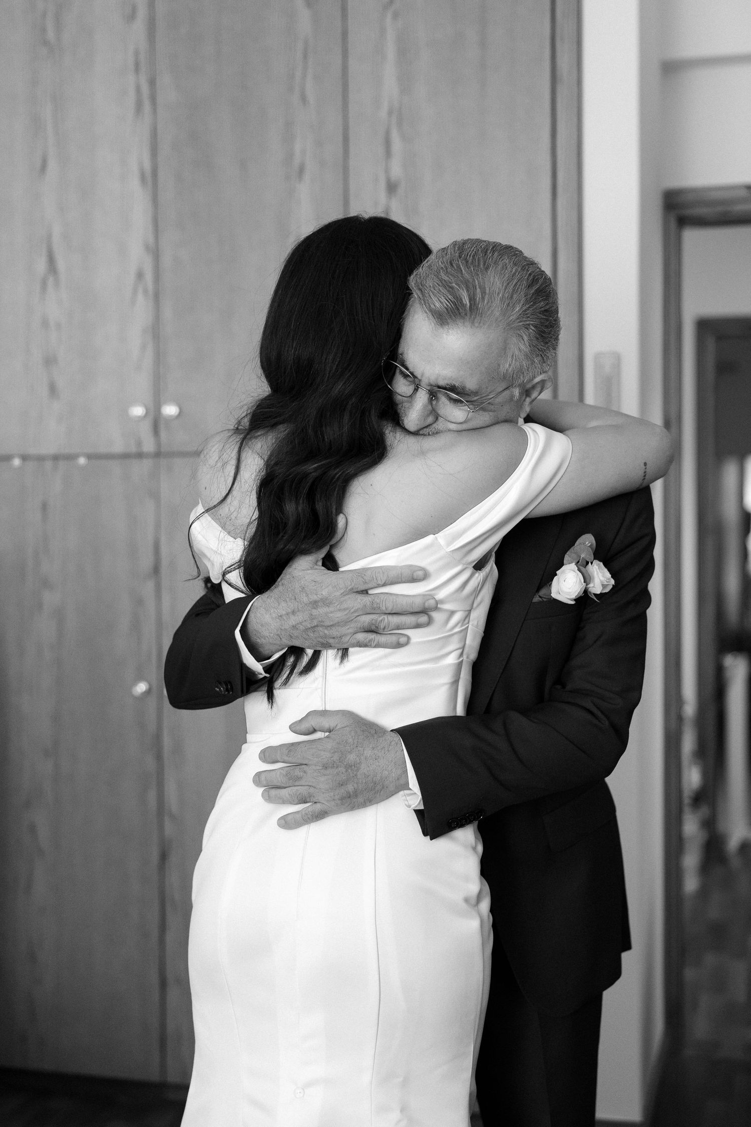 Black and white photo of a bride in a white gown sharing an emotional hug with an older man in a suit at a wedding.