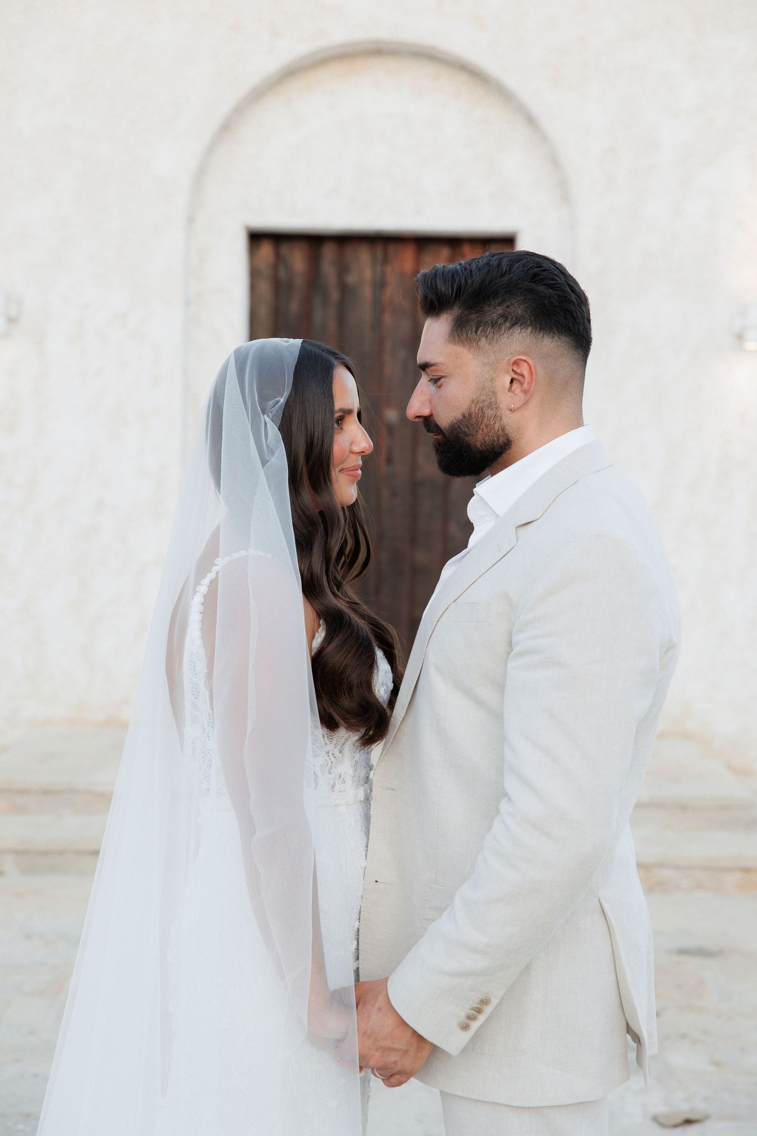 Bride and groom hold hands and gaze into each other eyes standing before a white stone archway.