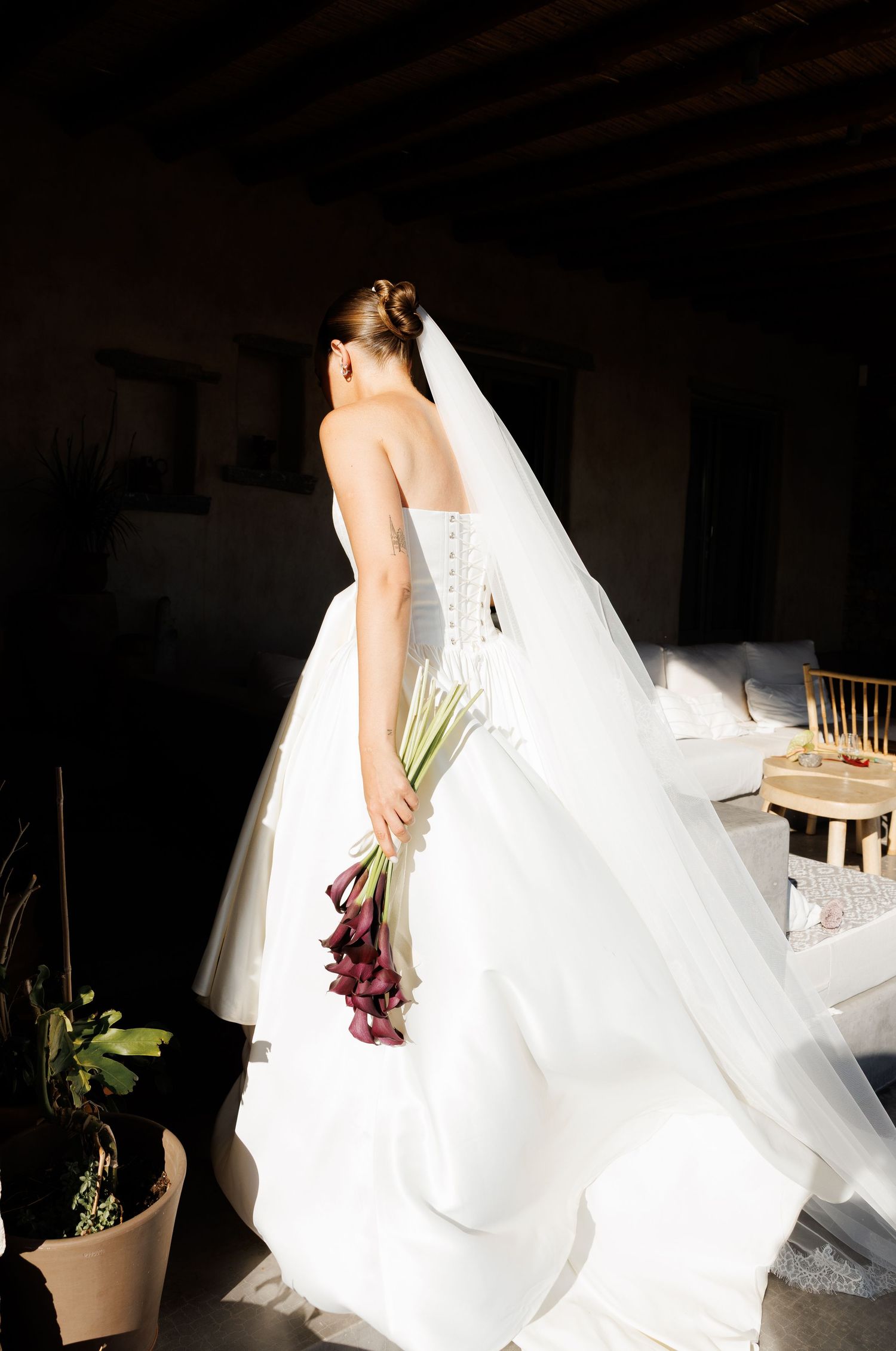 Bride in white strapless ballgown with veil holding purple calla lily bouquet standing in sunlit doorway.