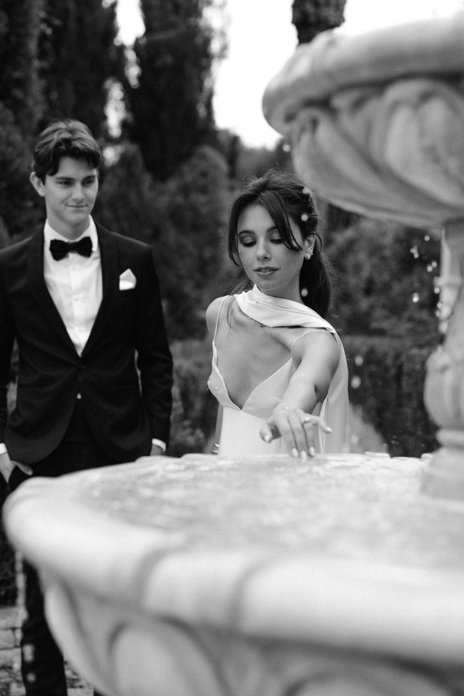 A black and white wedding photo showing a bride and groom at an ornate stone fountain in an elegant garden setting.