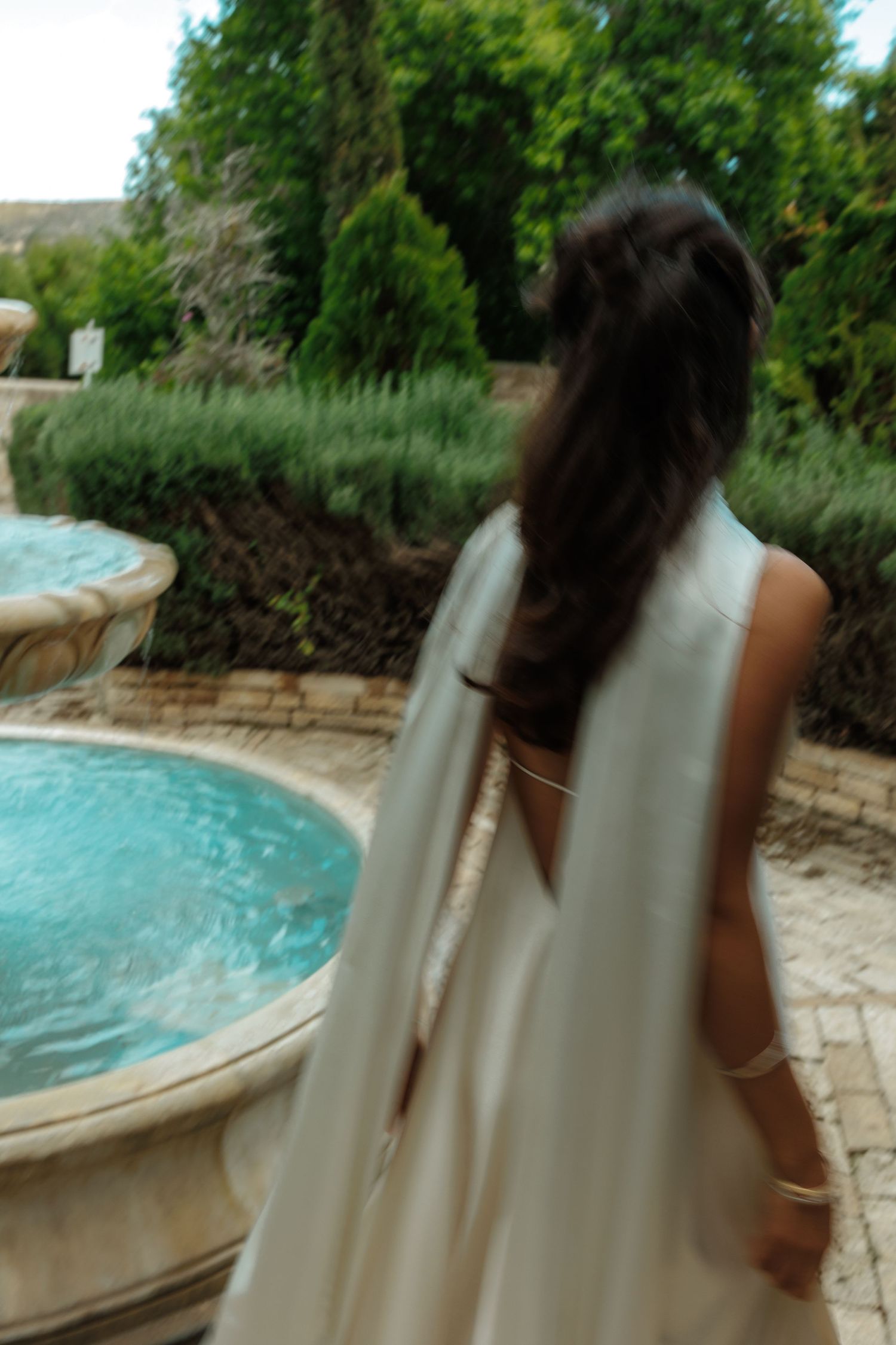 A blurry photo shows someone in a flowing white dress standing by an ornate garden pool with lush greenery in background.