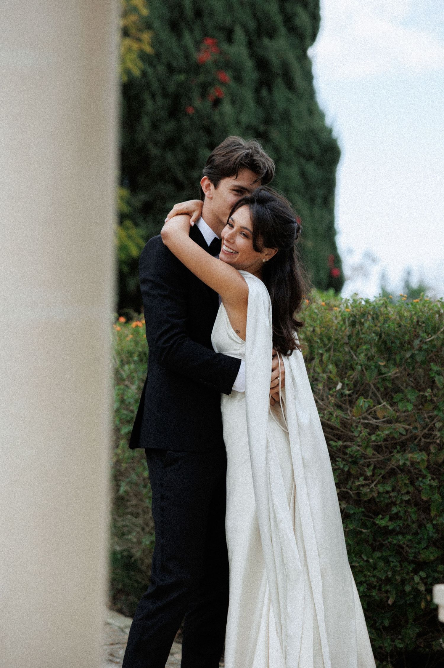 A tender embrace between newlyweds in formal attire against a backdrop of cypress trees and garden landscaping.