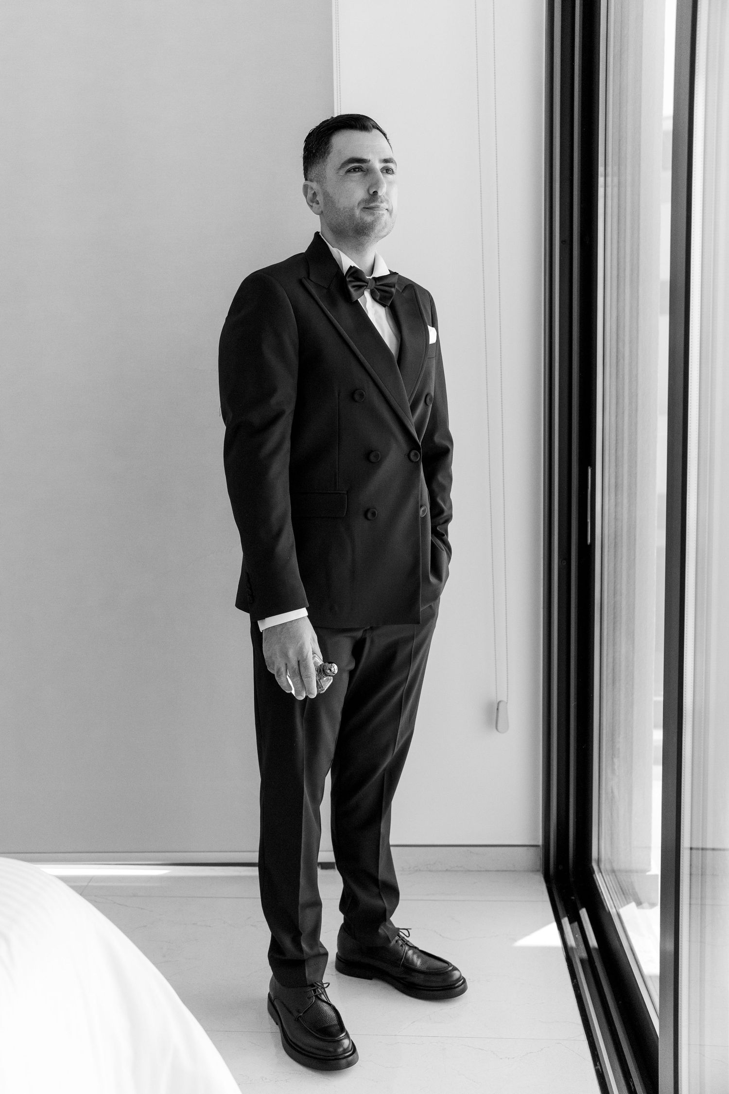 A black and white photo shows a groom in a classic double-breasted tuxedo standing by a window.