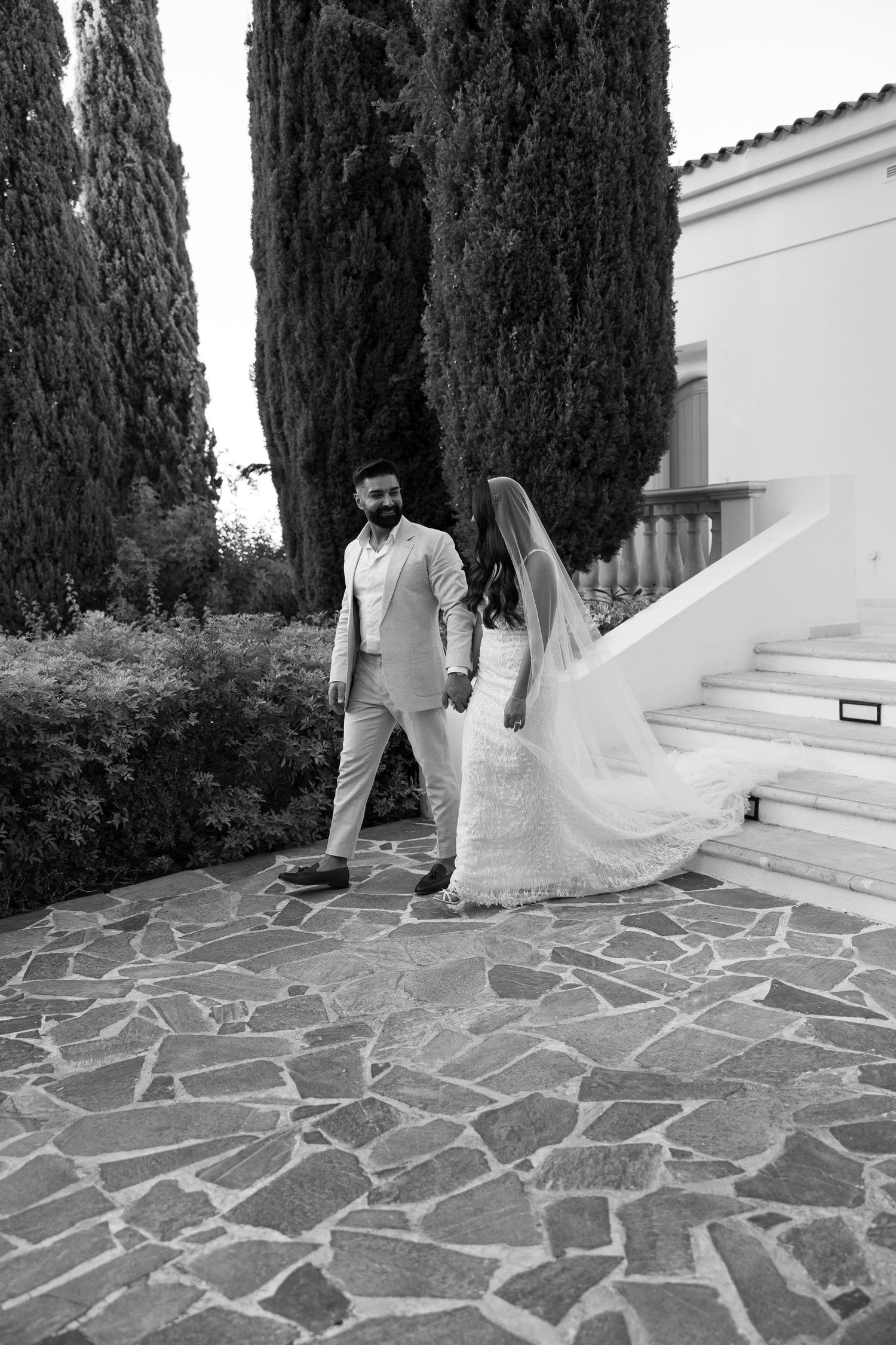 Black and white wedding photo of couple walking on stone path with cypress trees behind them.
